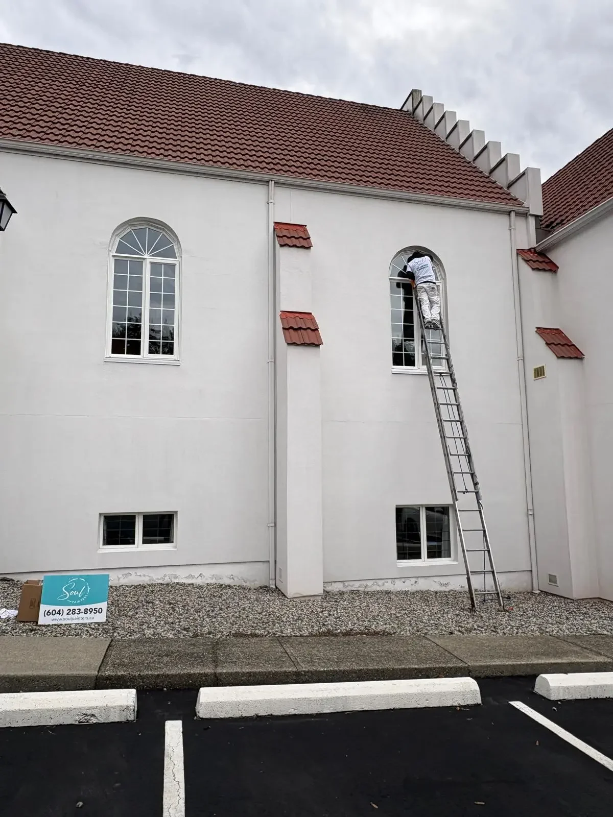 Painter on a ladder cleaning or repairing a tall arched window on a white building with red-tiled roof, offering free painting estimate and exterior painting services near you.