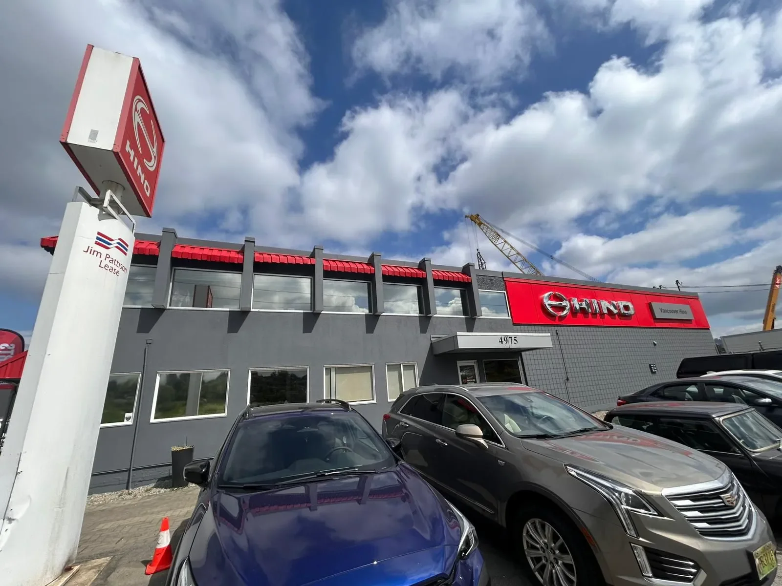 Exterior of a car dealership with various cars parked in front, a large sign with a logo, and a building with red and gray accents under a partly cloudy sky.