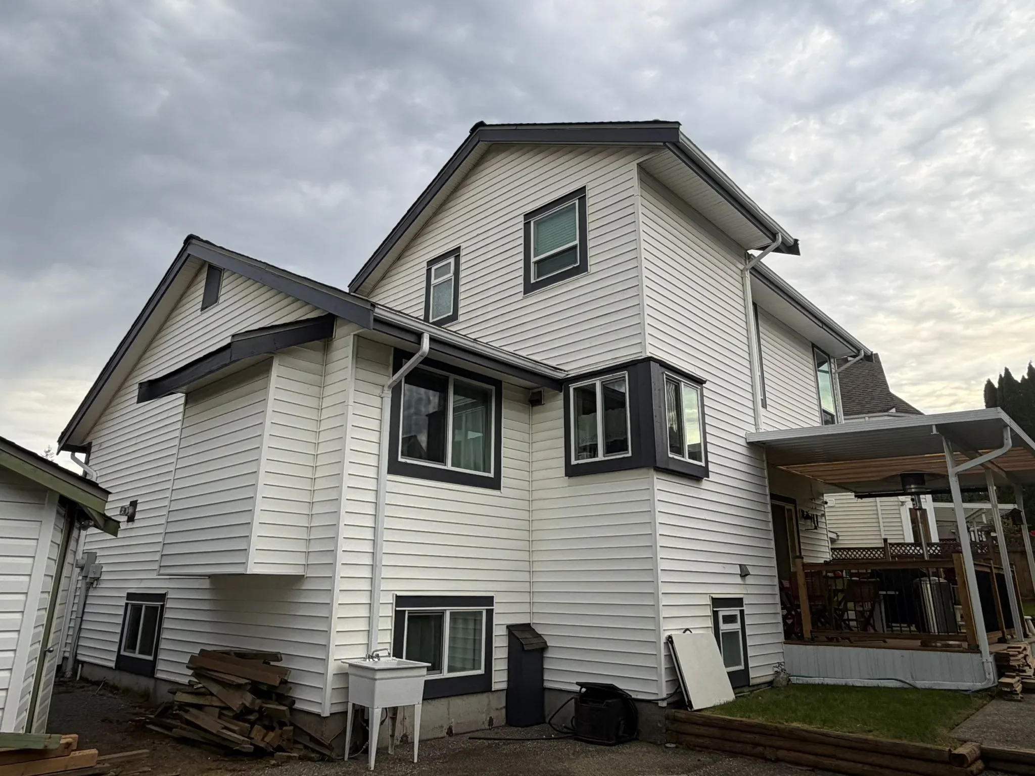 Rear view of a multi-story house with beige siding in North Vancouver, multiple windows, and a deck, under a cloudy sky.