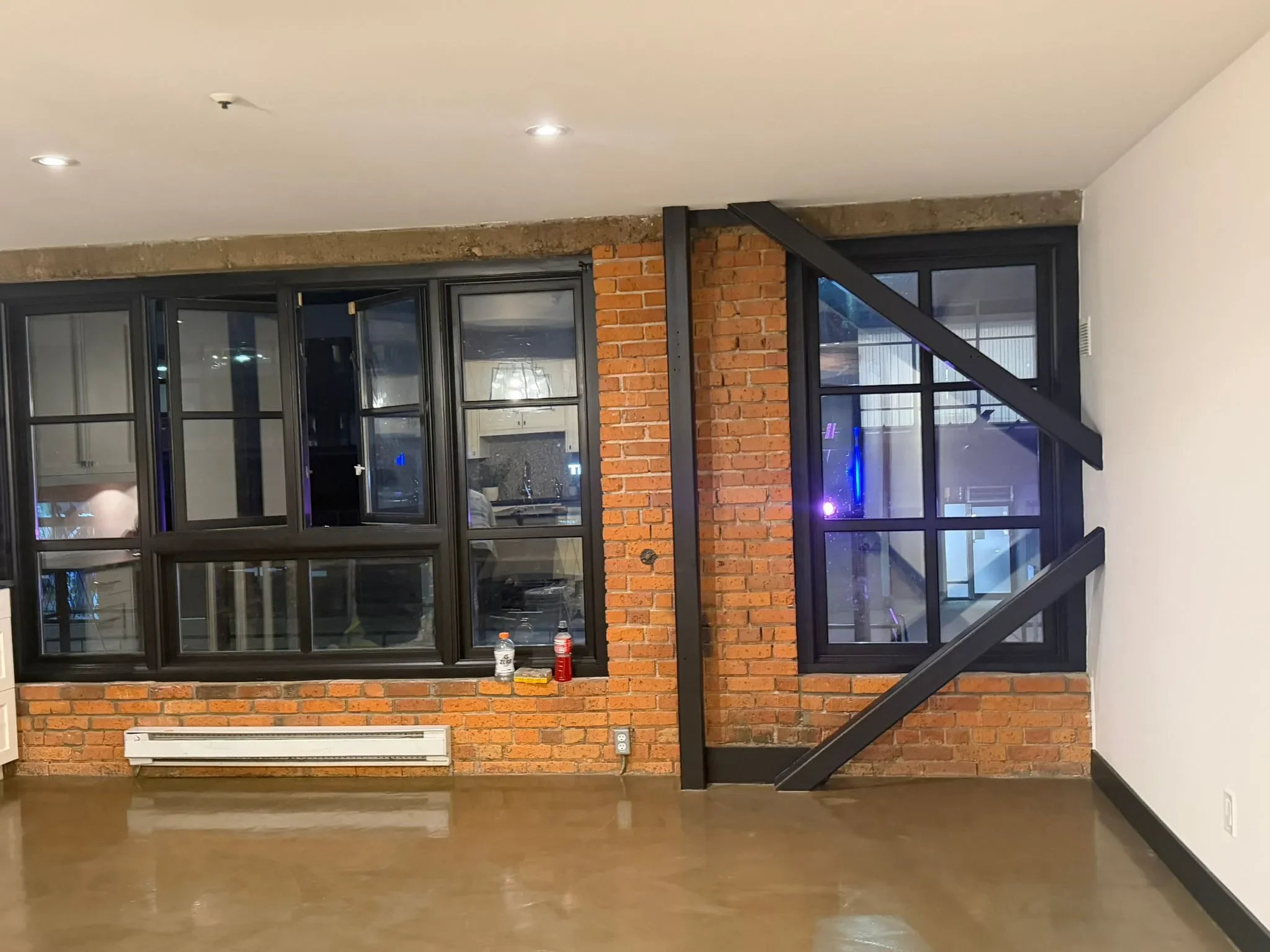 Interior of a modern building with a brick wall, large black-framed windows, a black staircase railing, a white wall, and a white electric heater on a polished concrete floor.