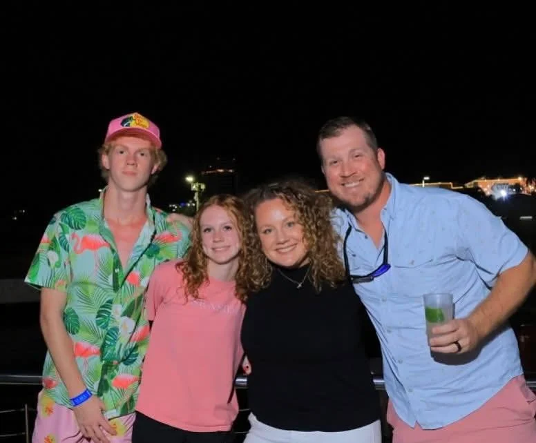 A group of four people standing outdoors at night, smiling and posing for a photo. The group includes two women and two young men, one of whom is holding a drink. The background features city lights and dark sky.