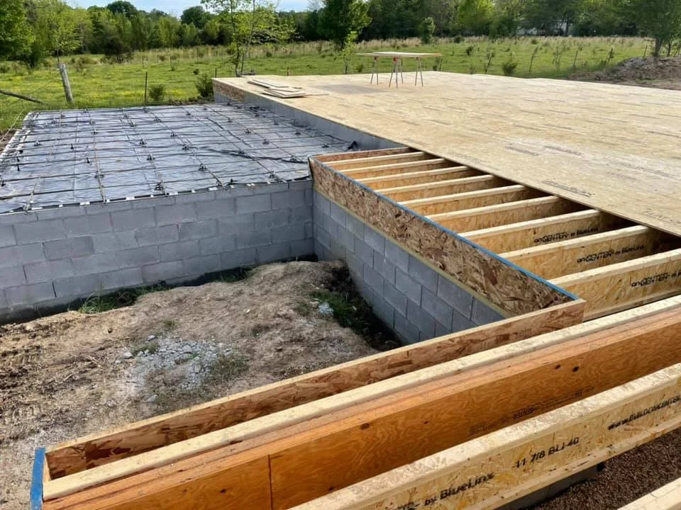 Construction site showing a partially built foundation with concrete blocks, a wooden frame, and rebar, surrounded by a grassy field.
