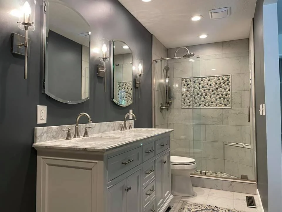 Bathroom with dual sinks, gray vanity, oval mirrors, wall sconces, walk-in shower with pebble tile accent, toilet, and modern fixtures.
