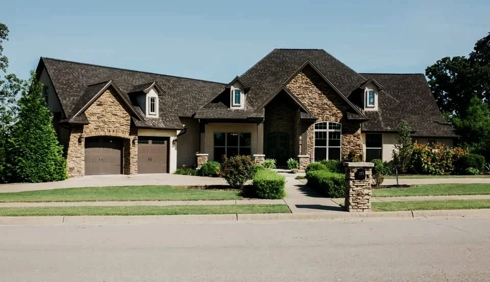 A large two-story house with a stone and stucco exterior, dark roof, and multiple dormer windows, surrounded by well-maintained lawn and bushes, with a sidewalk and street in front.