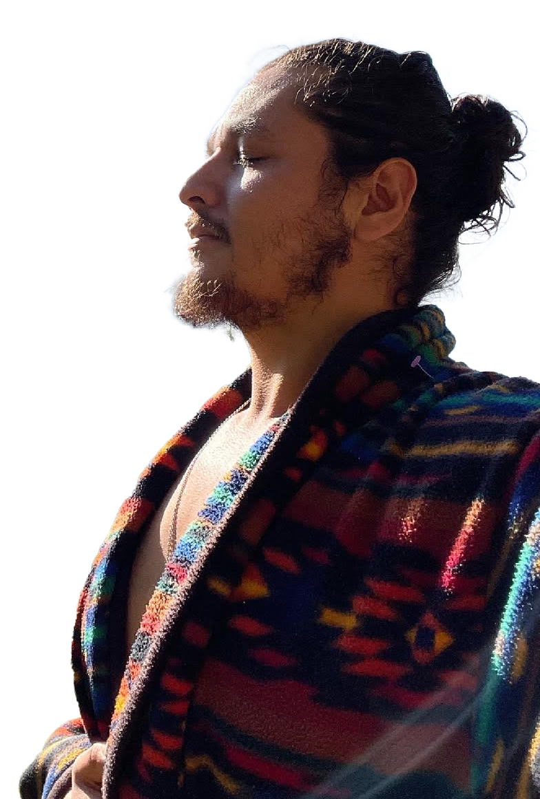 A man with long hair tied back, facial hair, and closed eyes, wearing a colorful patterned jacket, stands outdoors with sunlight shining on his face against a clear blue sky.