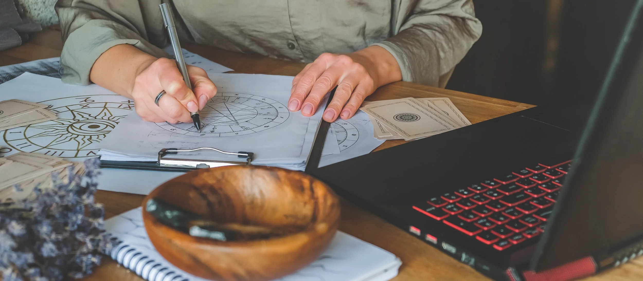 Person working on a detailed astrological or mystical chart with a black pen, surrounded by tarot cards, a wooden bowl, and a laptop on a wooden table.