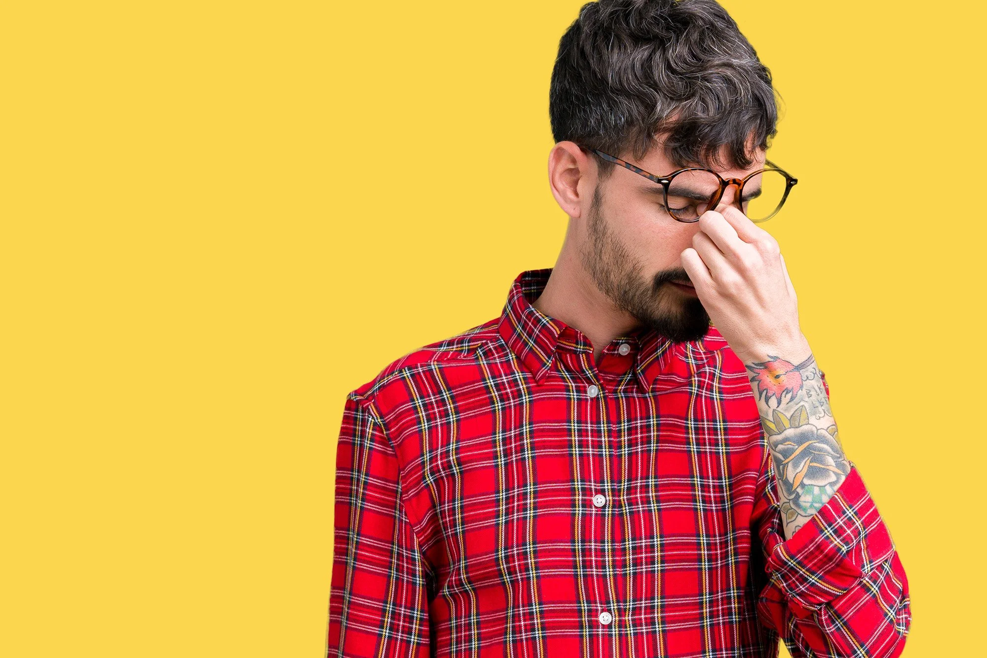 A man wearing glasses and a red plaid shirt stands against a yellow background, pinching the bridge of his nose with his right hand, with his eyes closed in apparent frustration or stress.