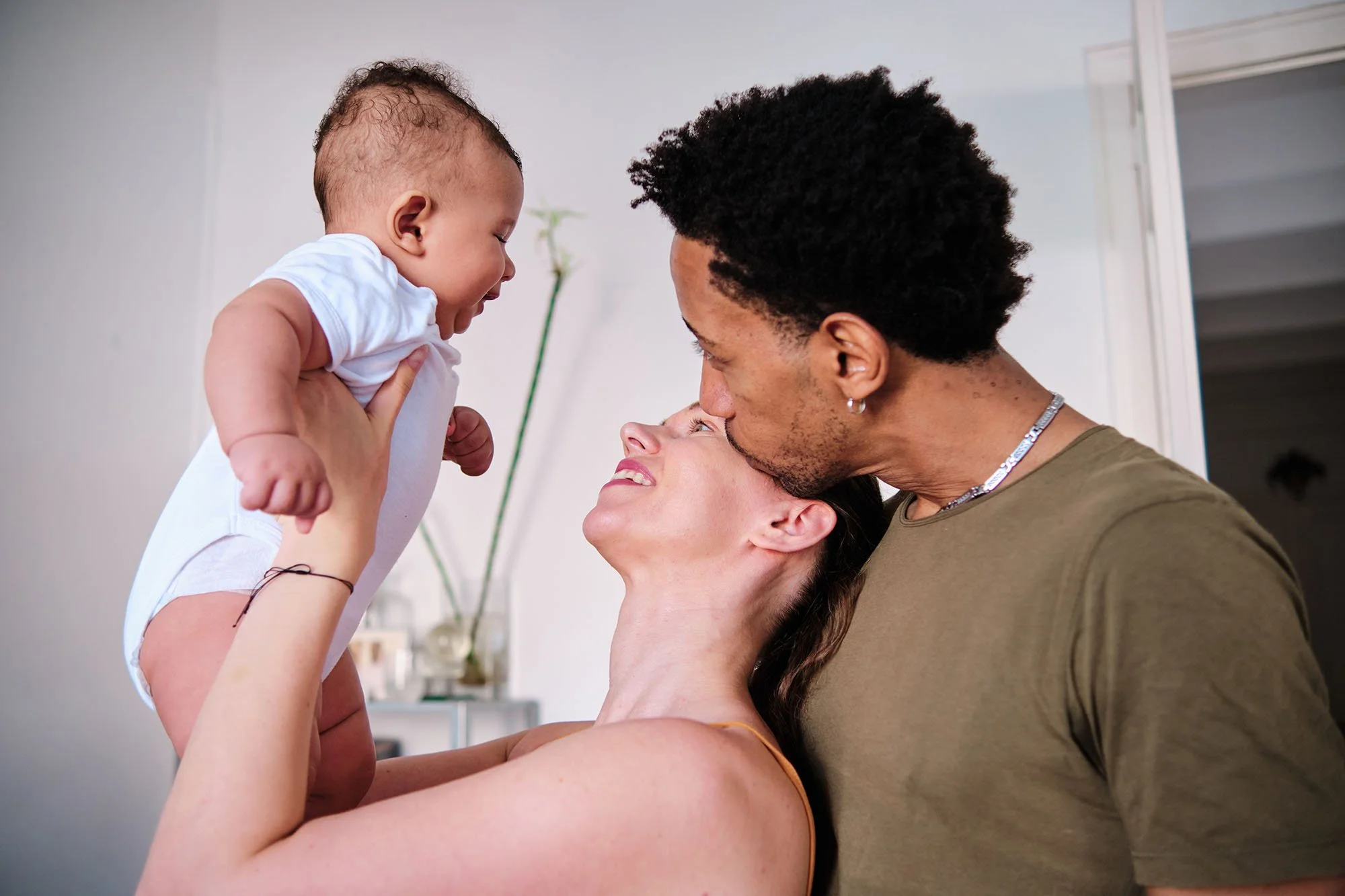 Family of three with a woman holding a baby, a man leaning over, lovingly looking at the baby, all smiling indoors.