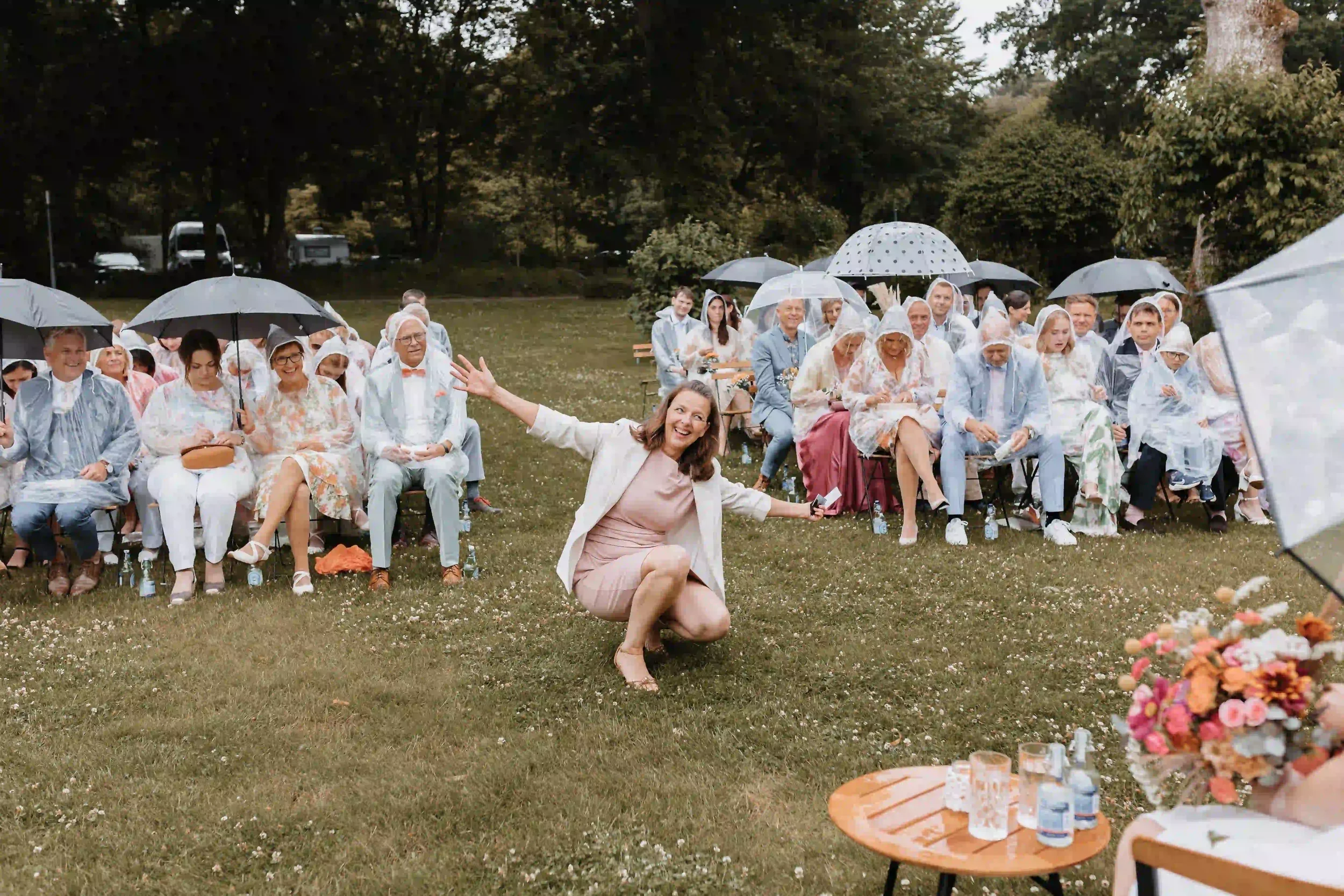 Rednerin hockt vor dem Publikum mit Regenschirmen bei einer freien Hochzeit im Regen