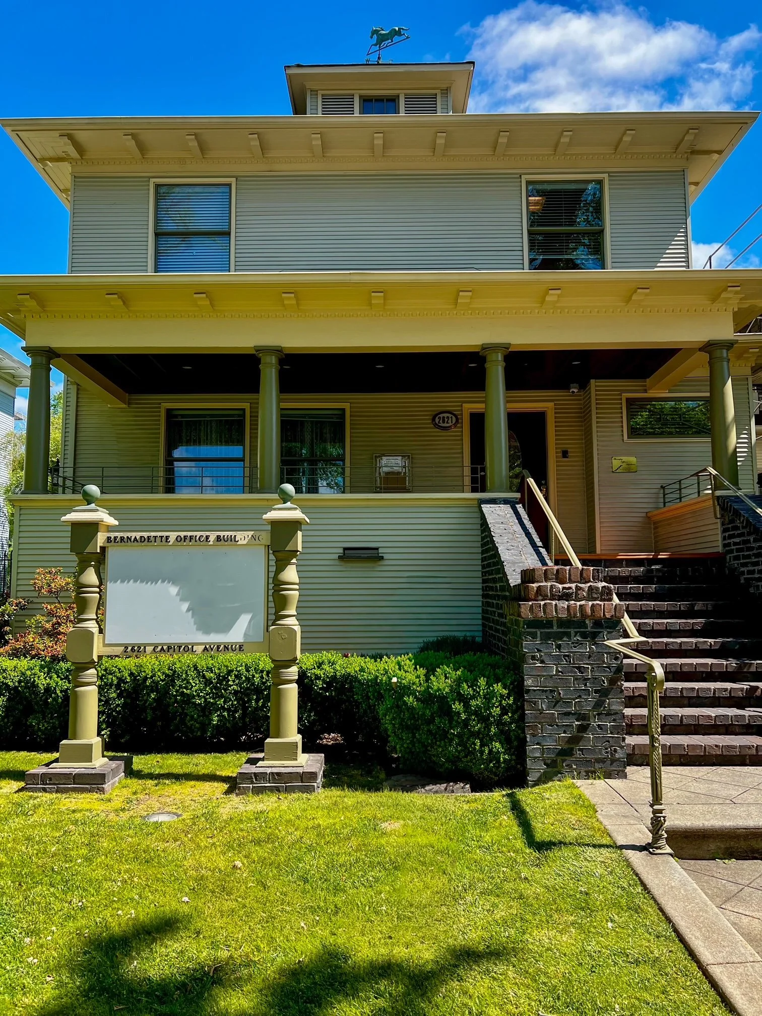 A three-story house with a porch, green columns, and a weather vane on the roof. It has a sign in front that reads 'Bernadette Office Building, 2621 Capitol Avenue.' The yard has green grass and shrubs, with a staircase leading up to the porch.