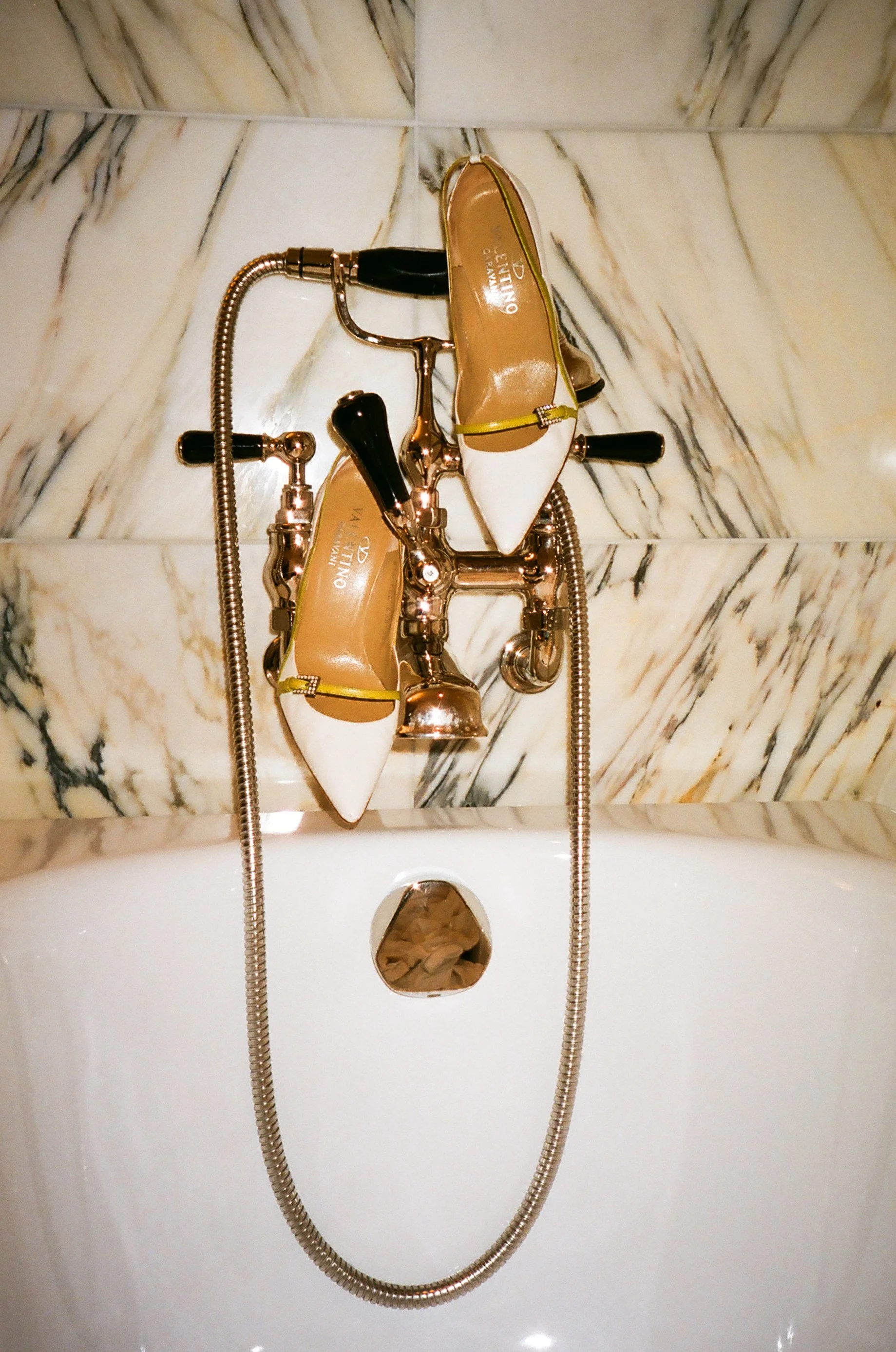 A bathtub faucet with a pair of white high-heeled shoes resting on top. The shoes have yellow straps and pointed toes. The faucet has a metallic finish and a flexible hose draped around it. Taken inside the Carlye Hotel.
