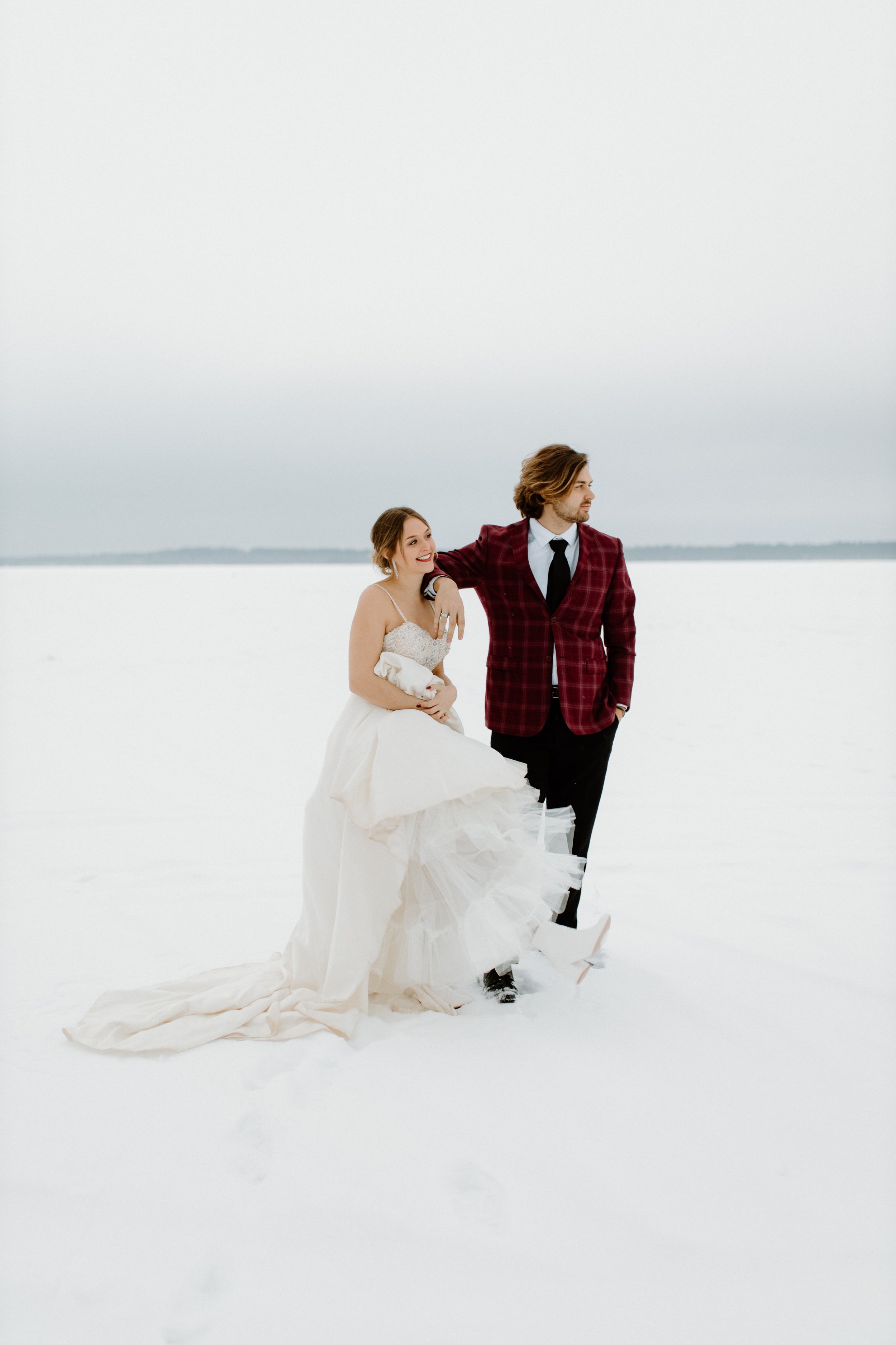 A couple dressed in wedding attire standing in a snowy landscape. The woman wears a white wedding gown and the man is in a dark red plaid suit with a black tie, and they are looking in different directions.