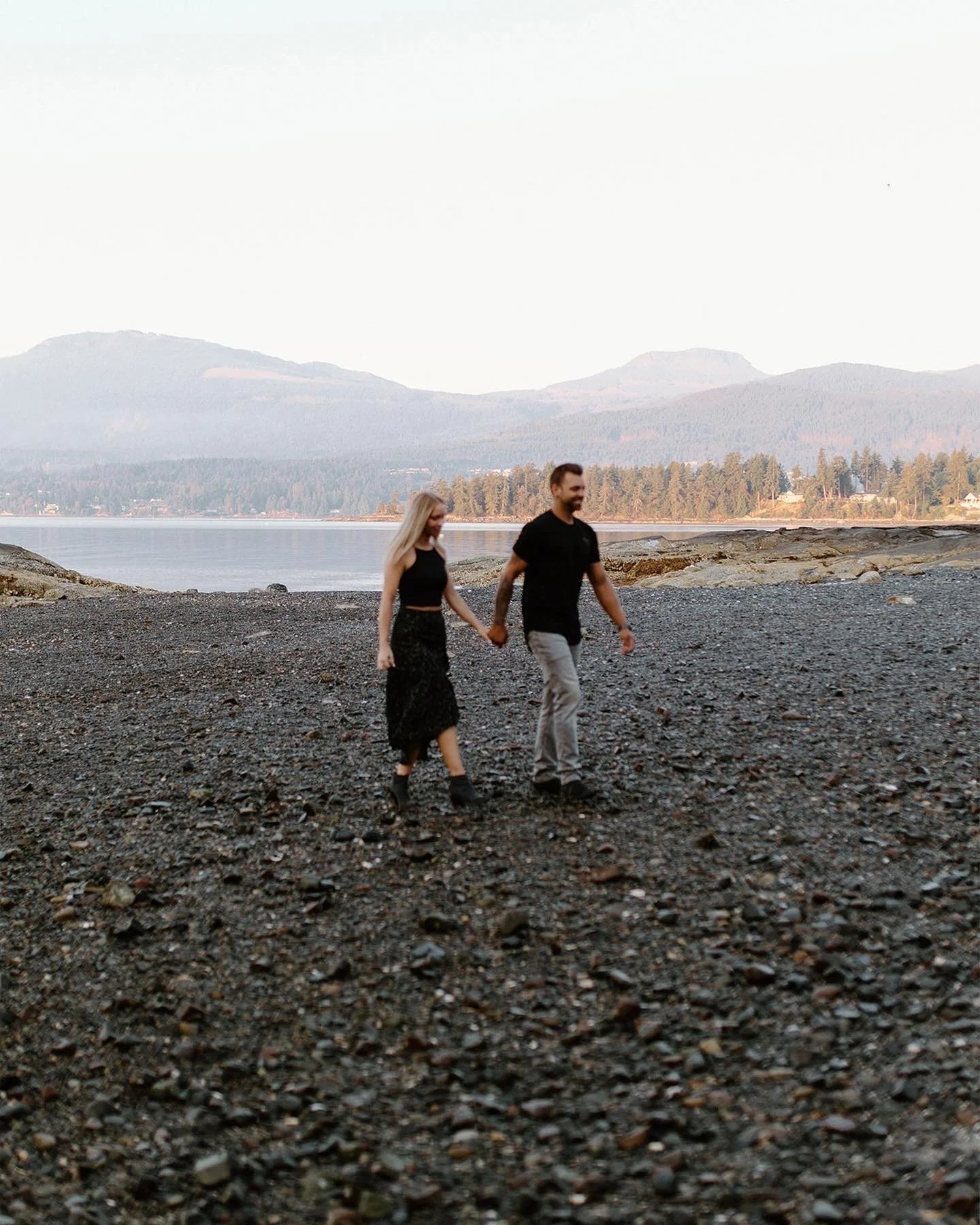 A couple holding hands and walking on a rocky beach with mountains and water in the background.