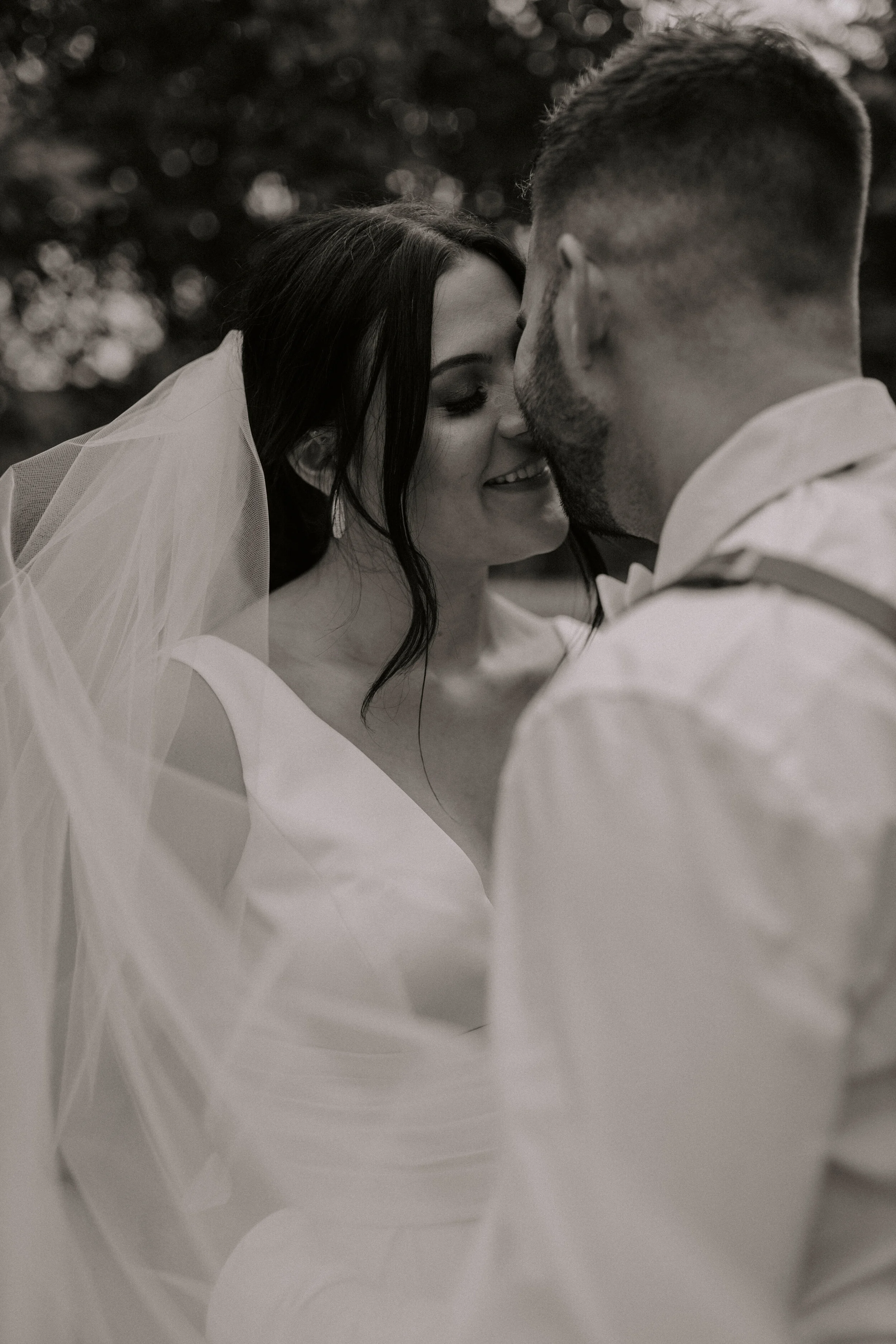 A black-and-white photo of a couple in wedding attire, nose to nose, smiling and looking into each other's eyes.