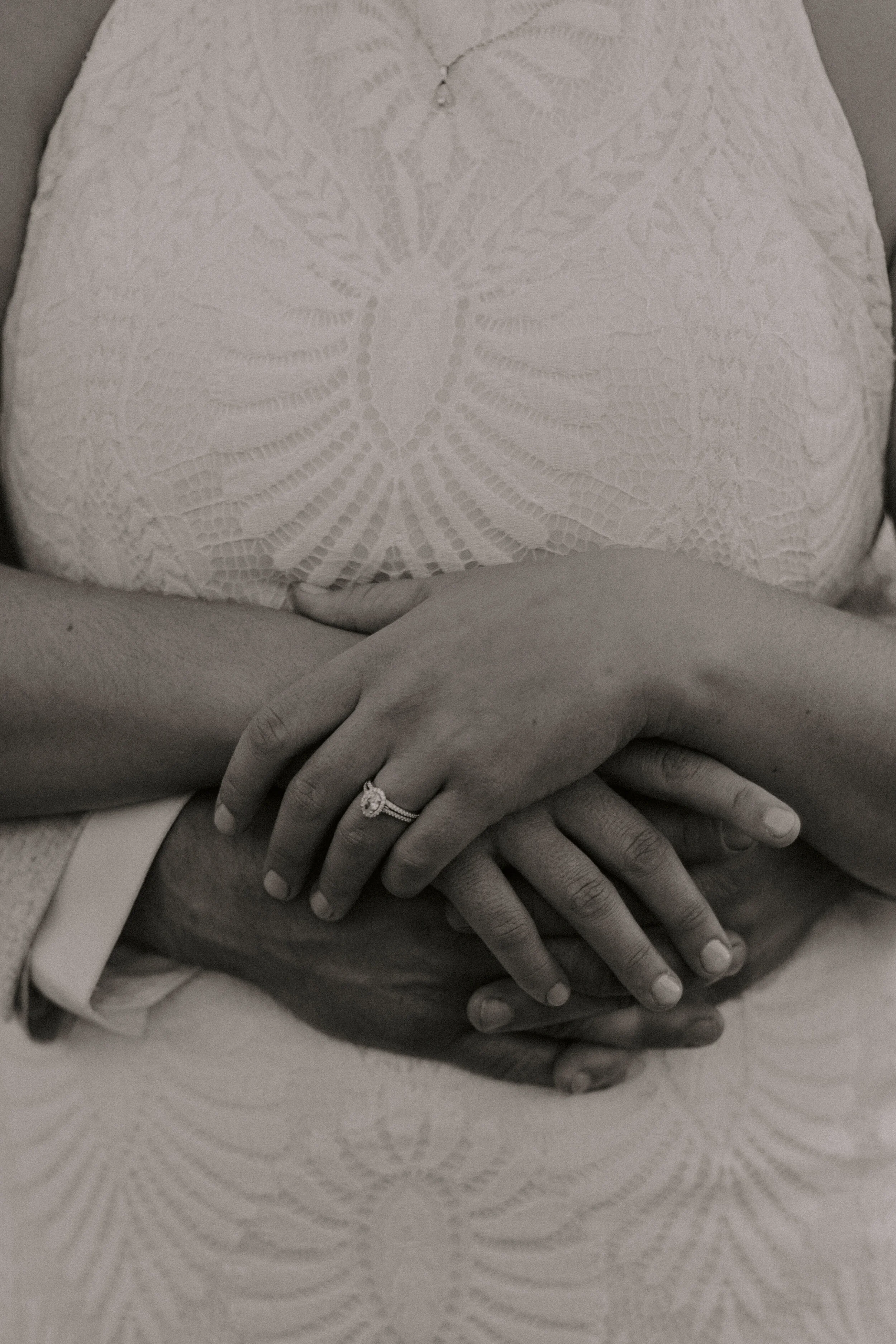Black and white photo of a couple's hands intertwined, with wedding rings, on fabric with a textured pattern.