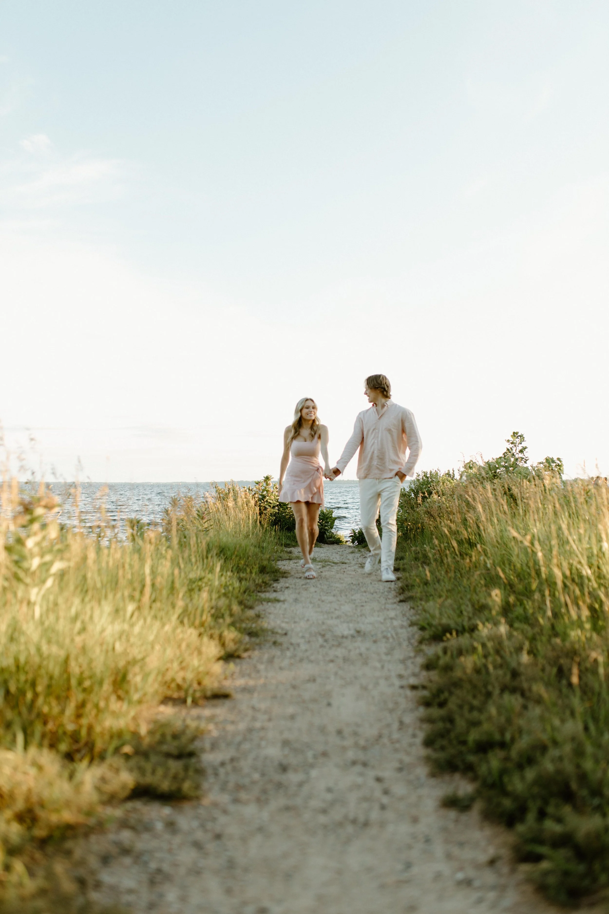 A young couple walking hand in hand along a nature trail by a lake, surrounded by tall grass and bushes, with a clear sky overhead.