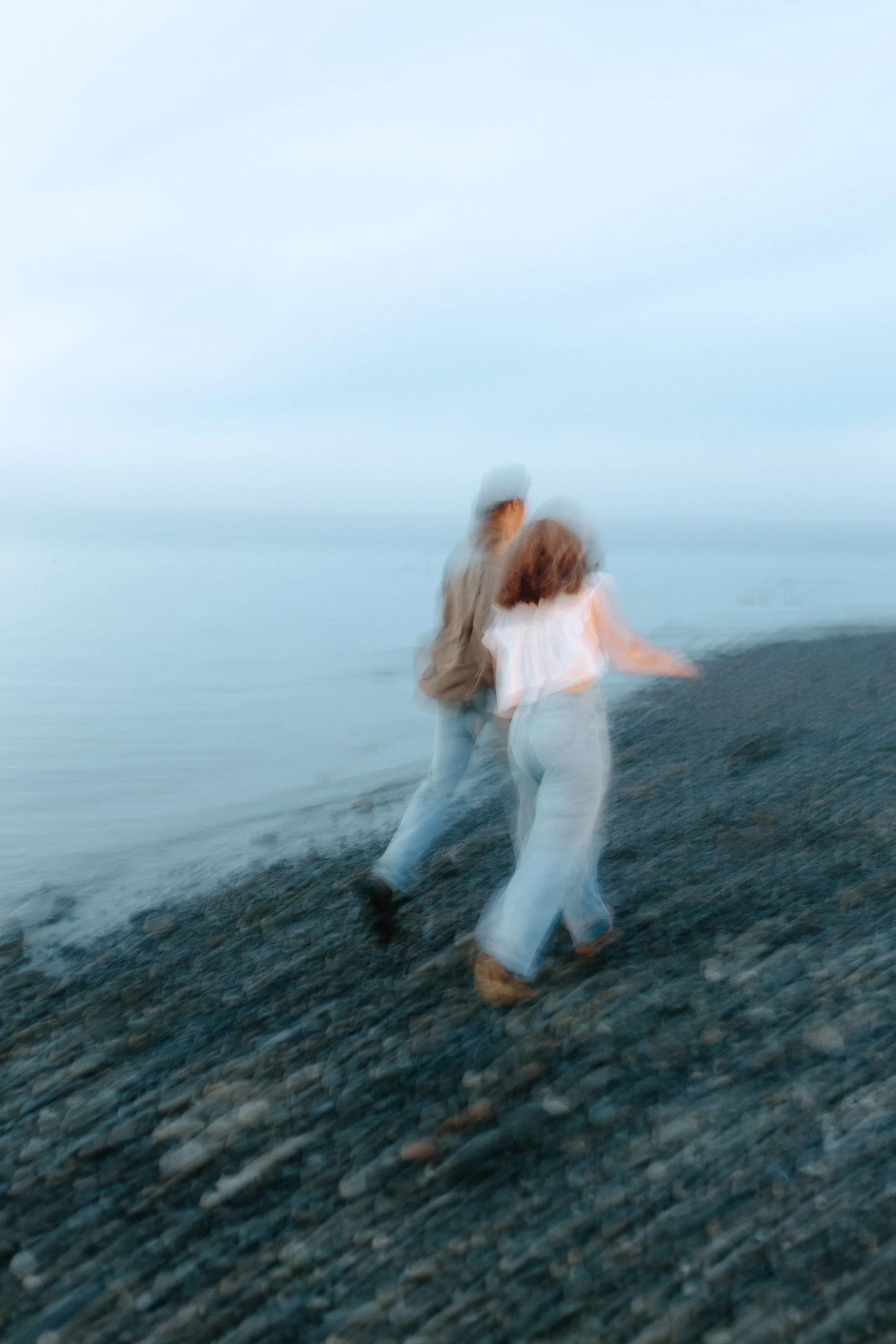 Blurred image of a couple walking on a pebbled beach near the water under an overcast sky.