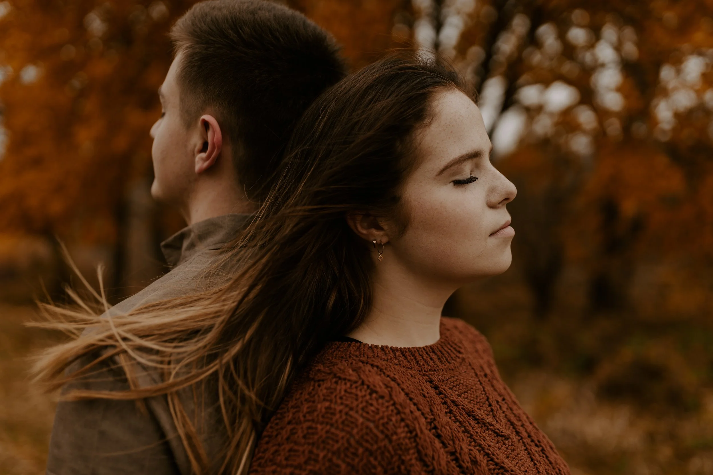 A young man and woman stand close with their backs to each other outdoors in autumn, with both having closed eyes and windswept hair, surrounded by orange and brown fall leaves.