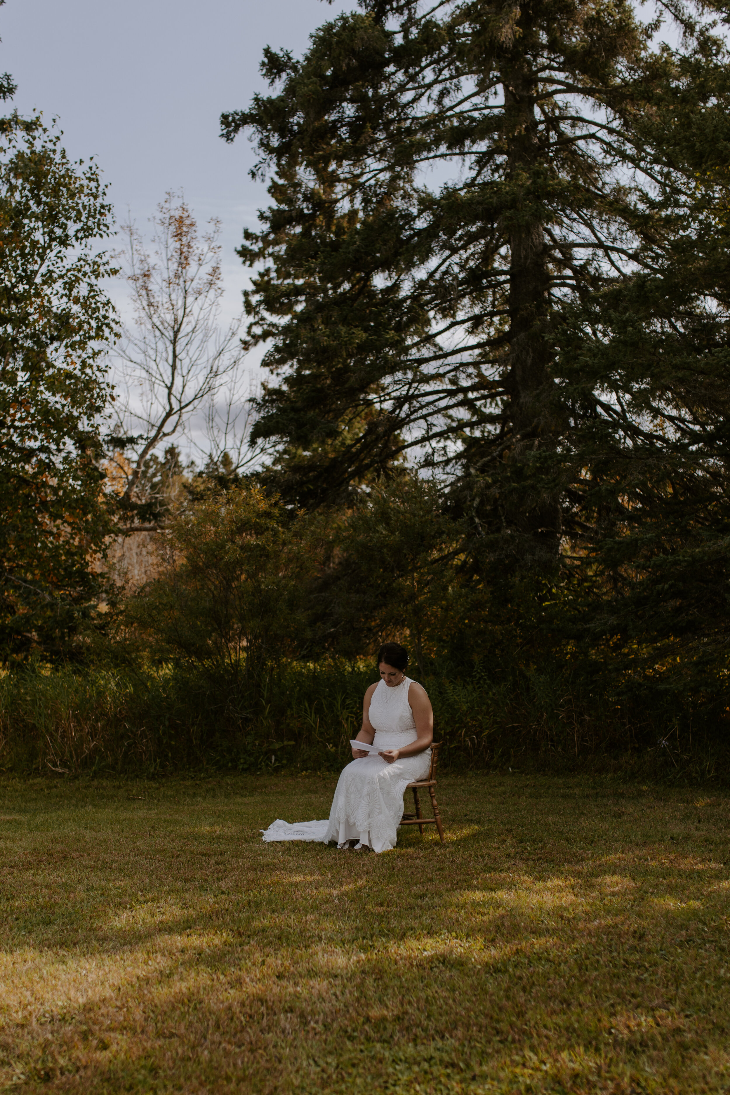 A woman in a white lace dress sits on a wooden chair on a grassy area, reading a book, with tall trees and shrubs in the background during sunset.