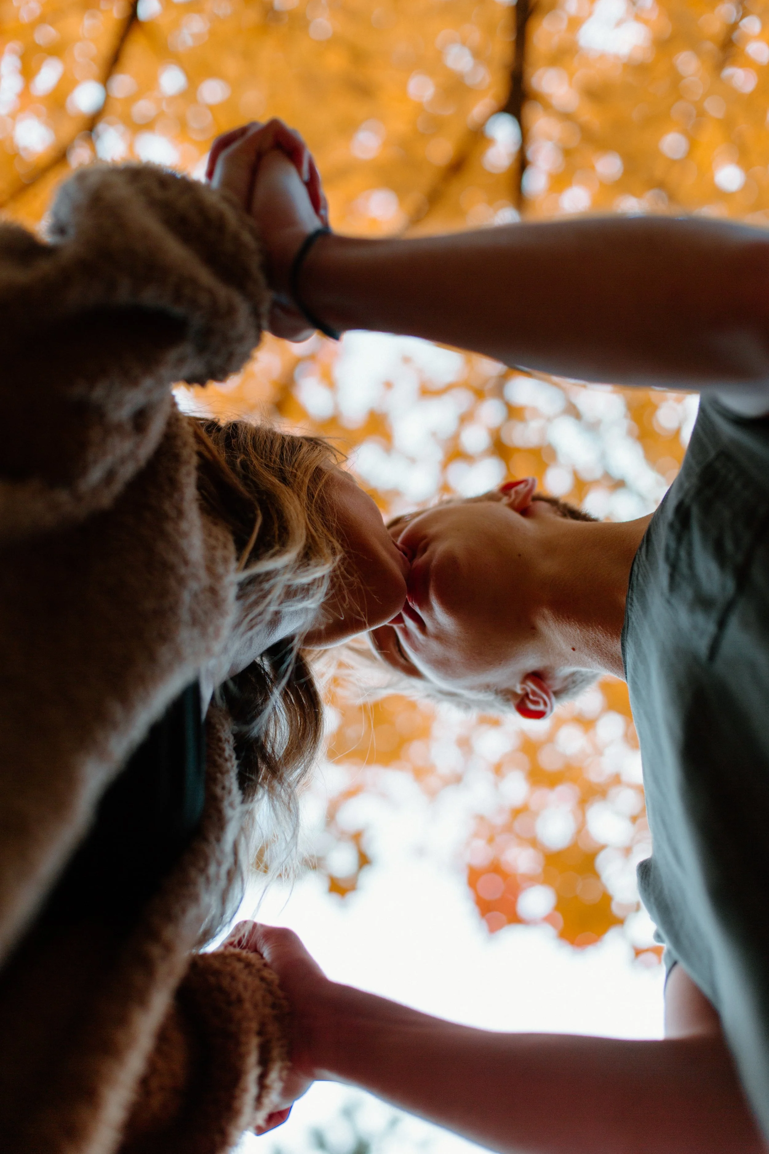 A couple sharing a kiss outdoors beneath orange autumn leaves, with the camera pointed upward capturing their faces and the leaves.