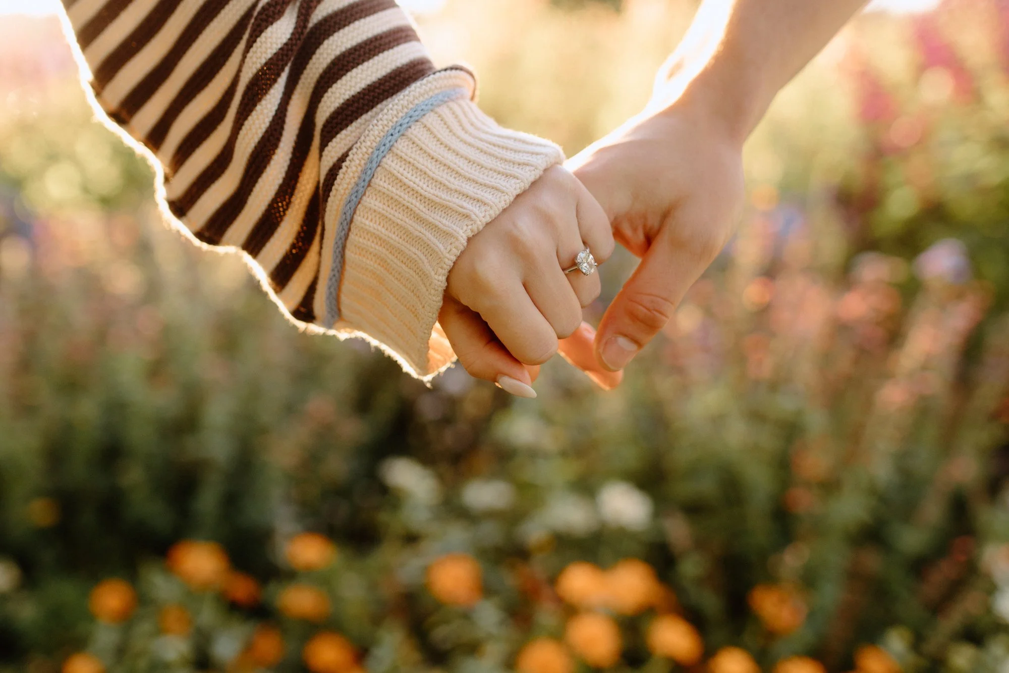 Close-up of two people holding hands, with one person's hand wearing a diamond ring, in an outdoor setting with blurred background of orange berries and greenery.
