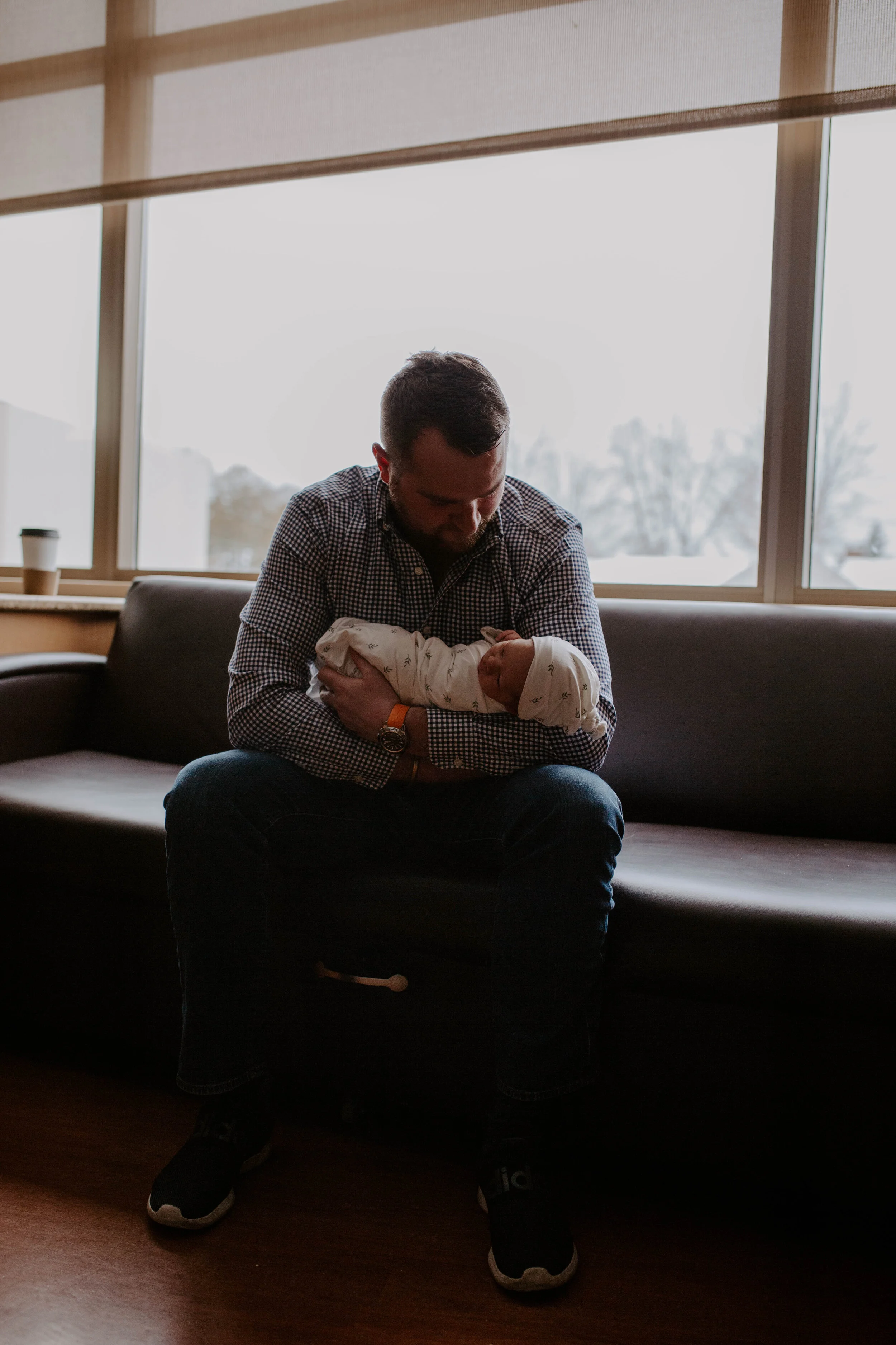 A man sitting on a black couch holding a baby with a white blanket, near large windows with a mountain view, during daytime.