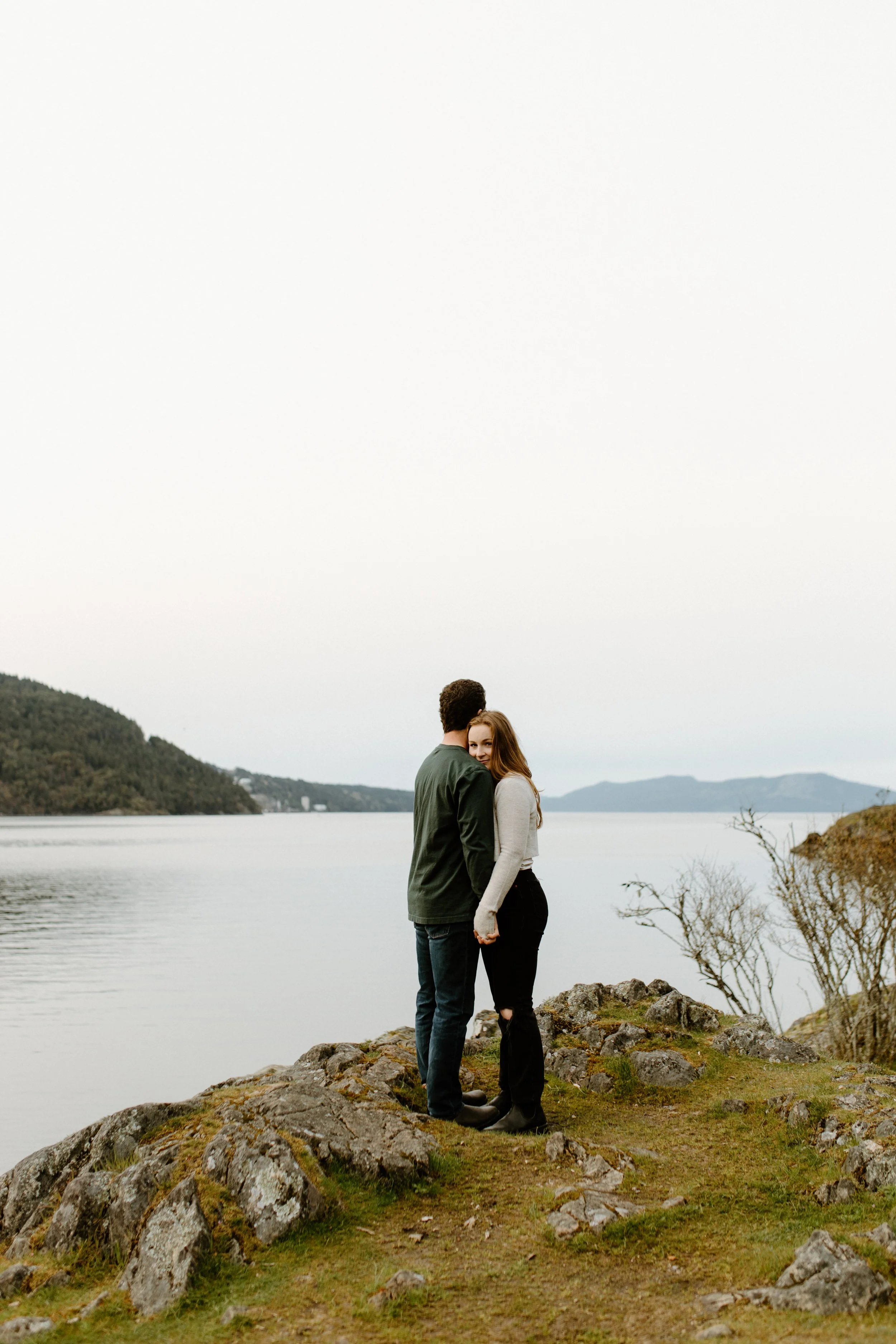 A young couple stands close together on a rocky shoreline near a body of water, embracing each other with lush green hills and a cloudy sky in the background.