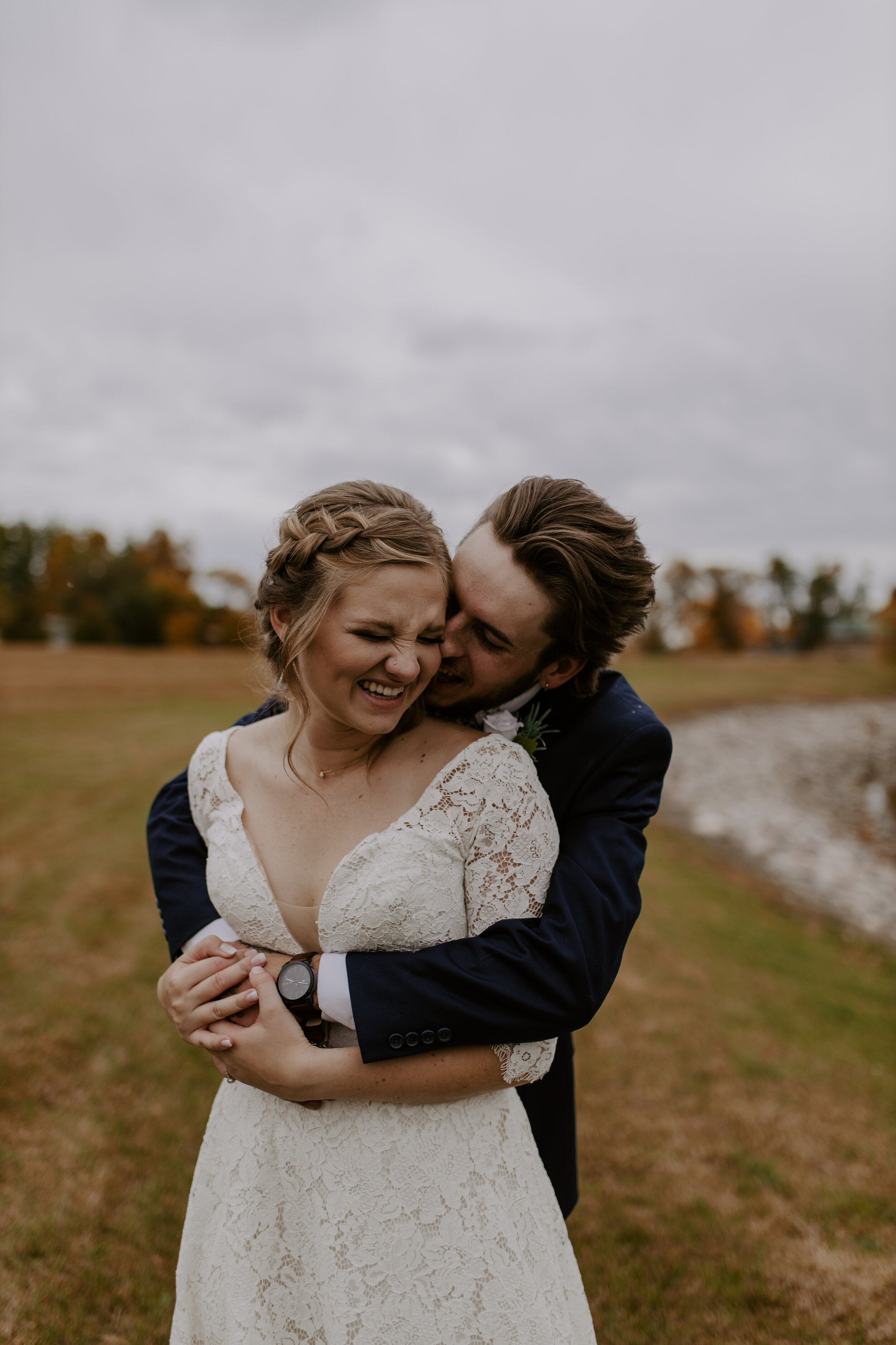 A bride and groom are embracing outdoors on a cloudy day, with fall trees in the background. The bride is smiling with eyes closed, wearing a lace wedding dress with a scoop neckline. The groom is behind her, smiling with his head close to hers, wear
