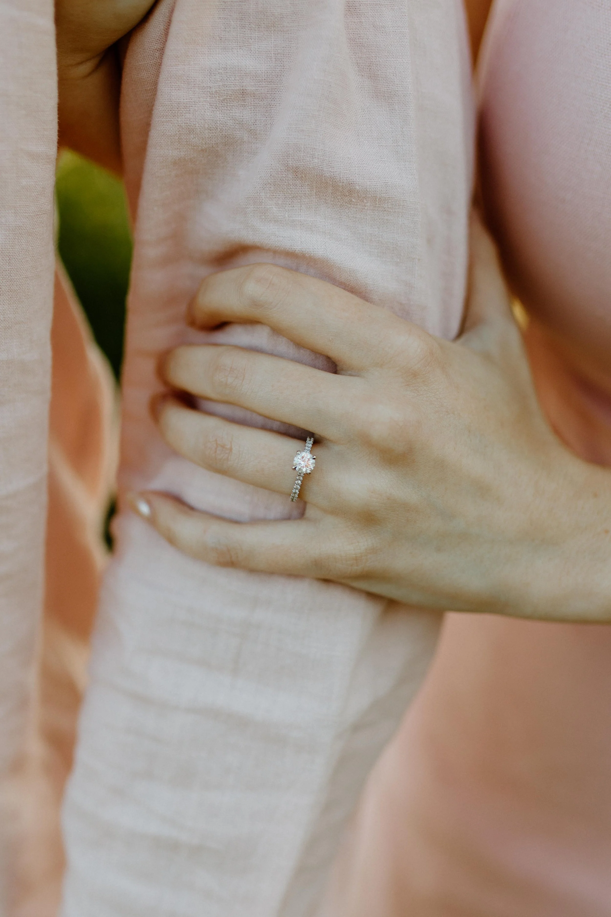 Close-up of a hand with a ring resting on the stomach area, wearing a light pink fabric shirt. The background is soft pink.