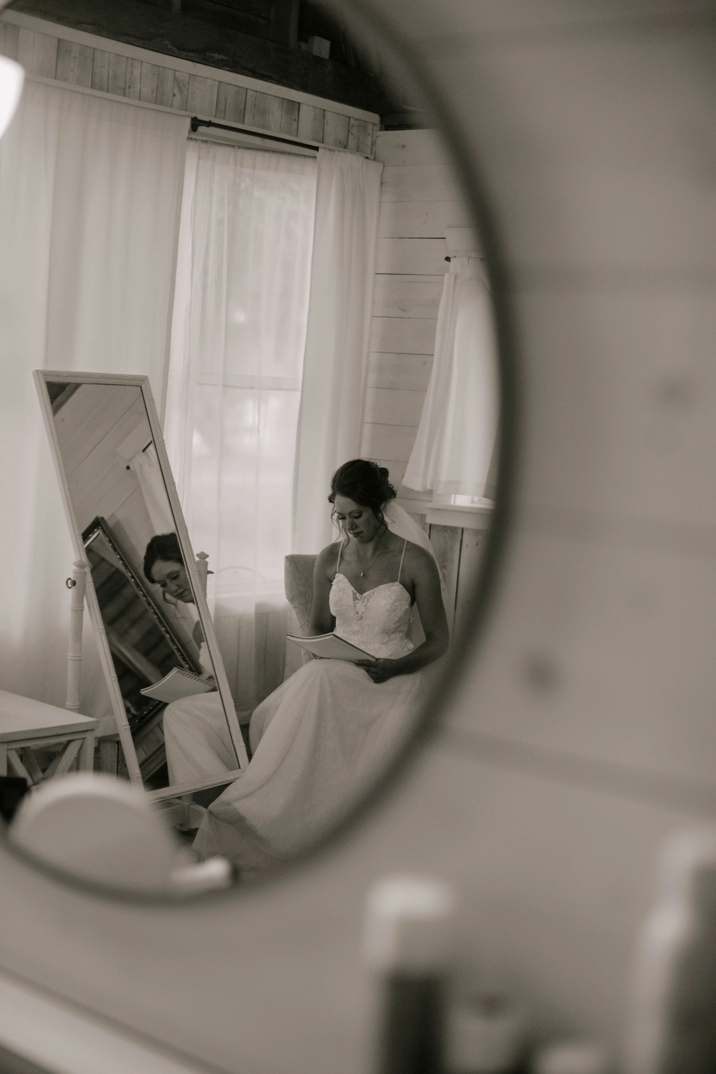 A woman in a wedding dress sitting in front of a mirror, captured through a reflection in a circular mirror.