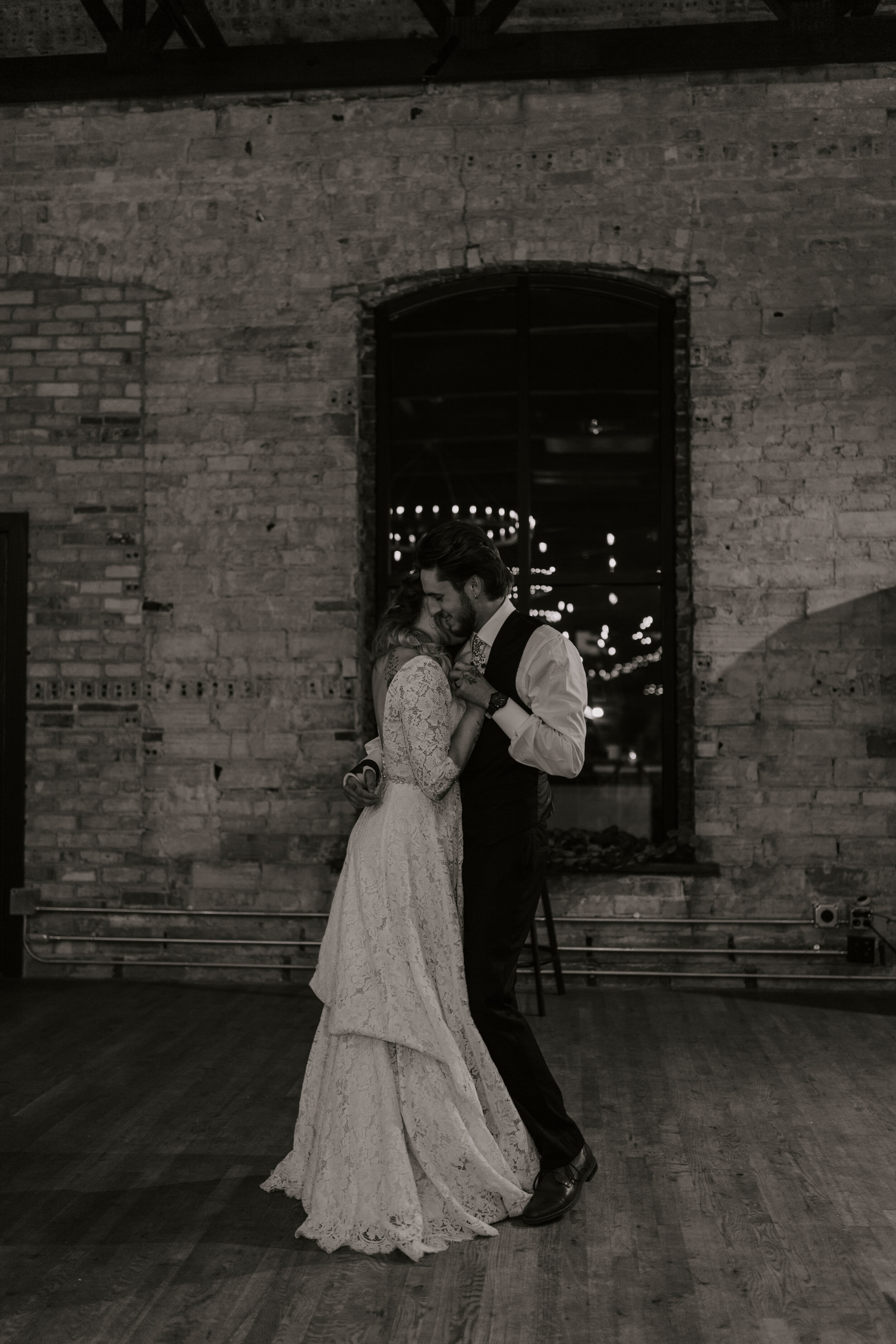 A couple sharing a dance in a rustic setting with exposed brick walls and string lights reflected in a window behind them.