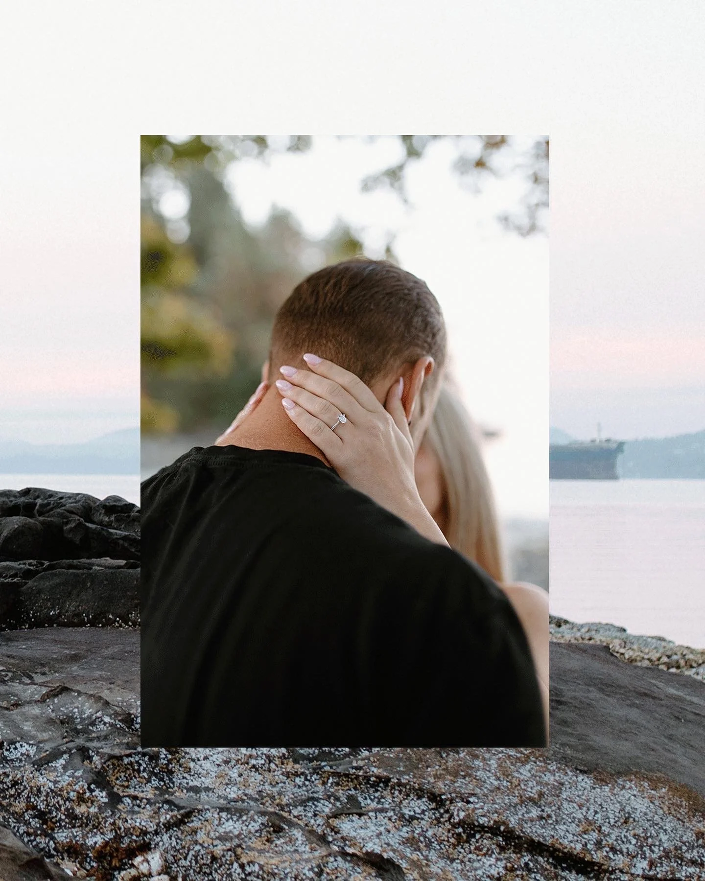 A couple embracing by the ocean, with the woman wearing an engagement ring, and the man facing away with short hair, seen through a cutout in a rocky shoreline.