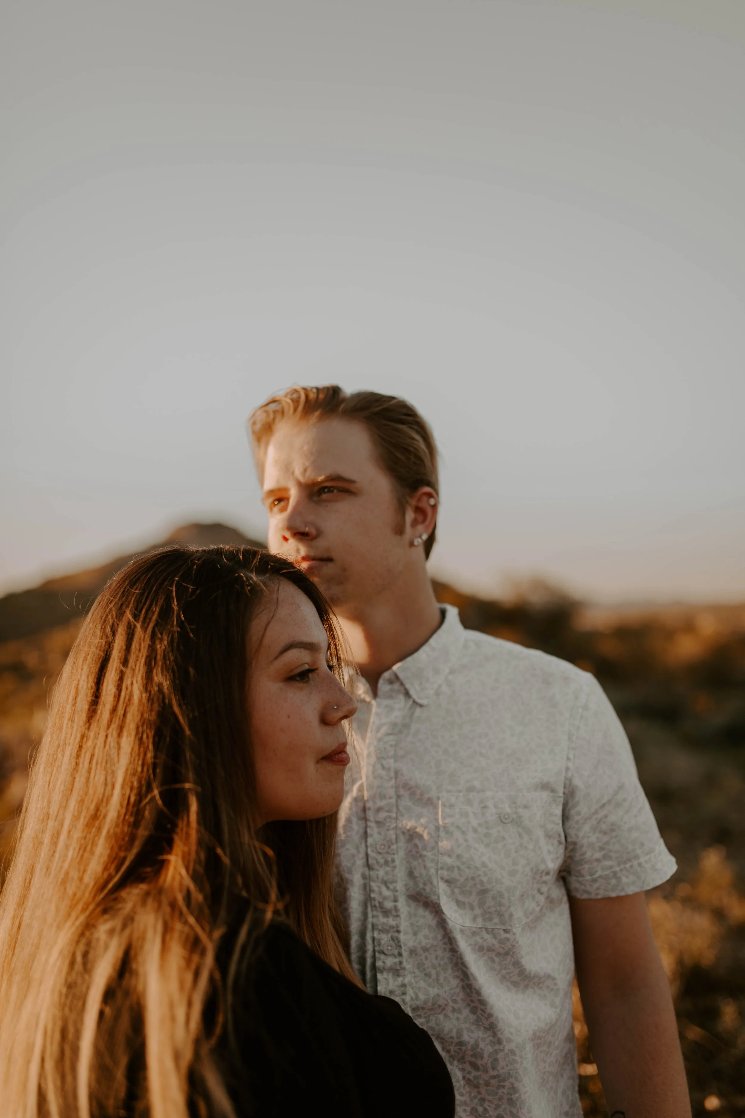 A young woman with long red hair and a young man with short light brown hair standing outdoors during sunset, with a mountainous landscape in the background.