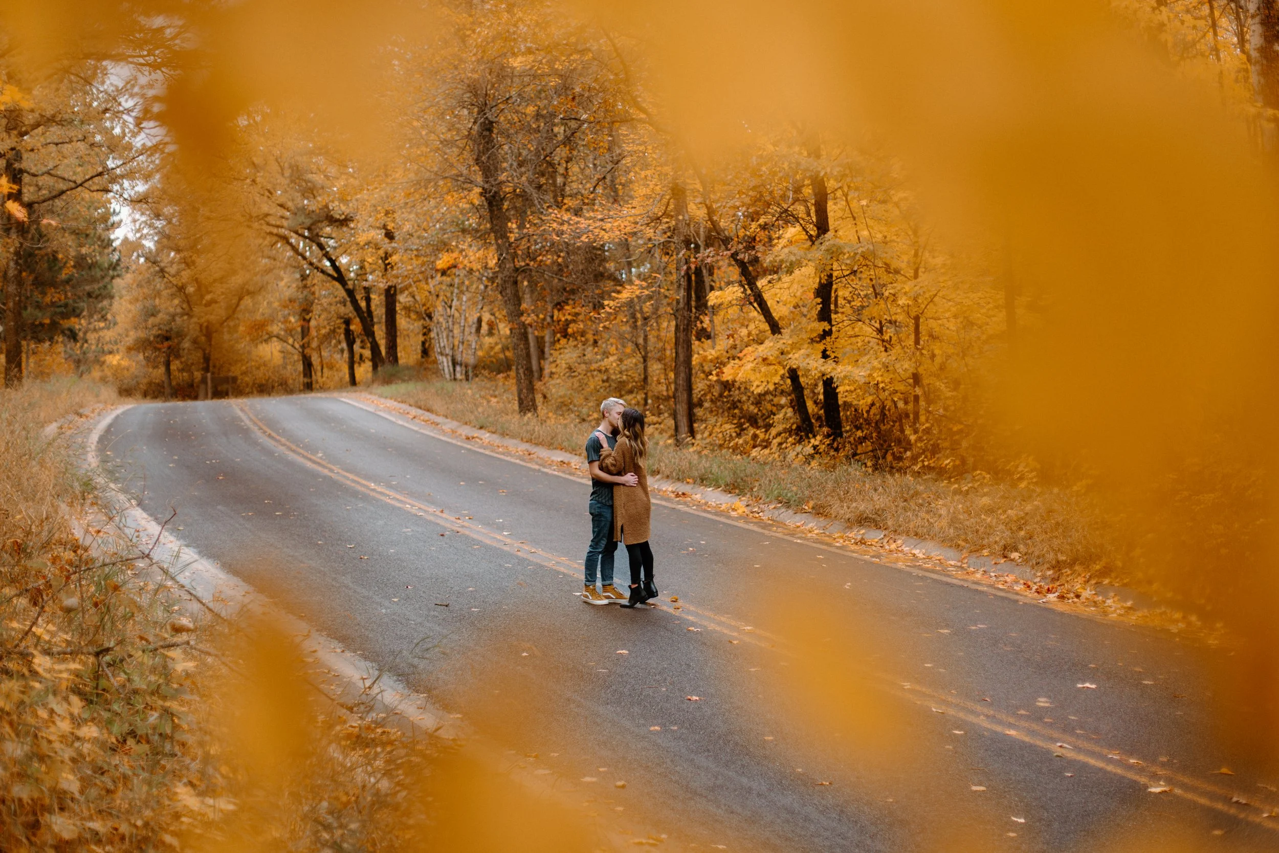 A couple embraces on a winding road surrounded by orange and yellow autumn foliage.