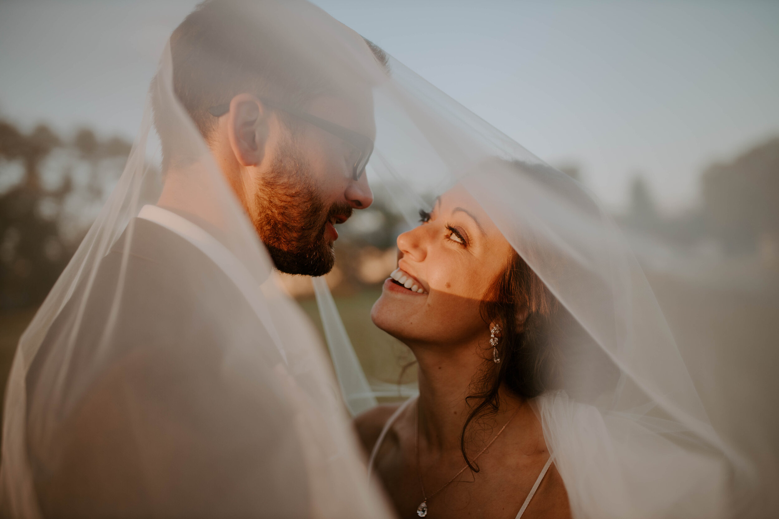 A bride and groom under a wedding veil, smiling and looking into each other's eyes outside during sunset.