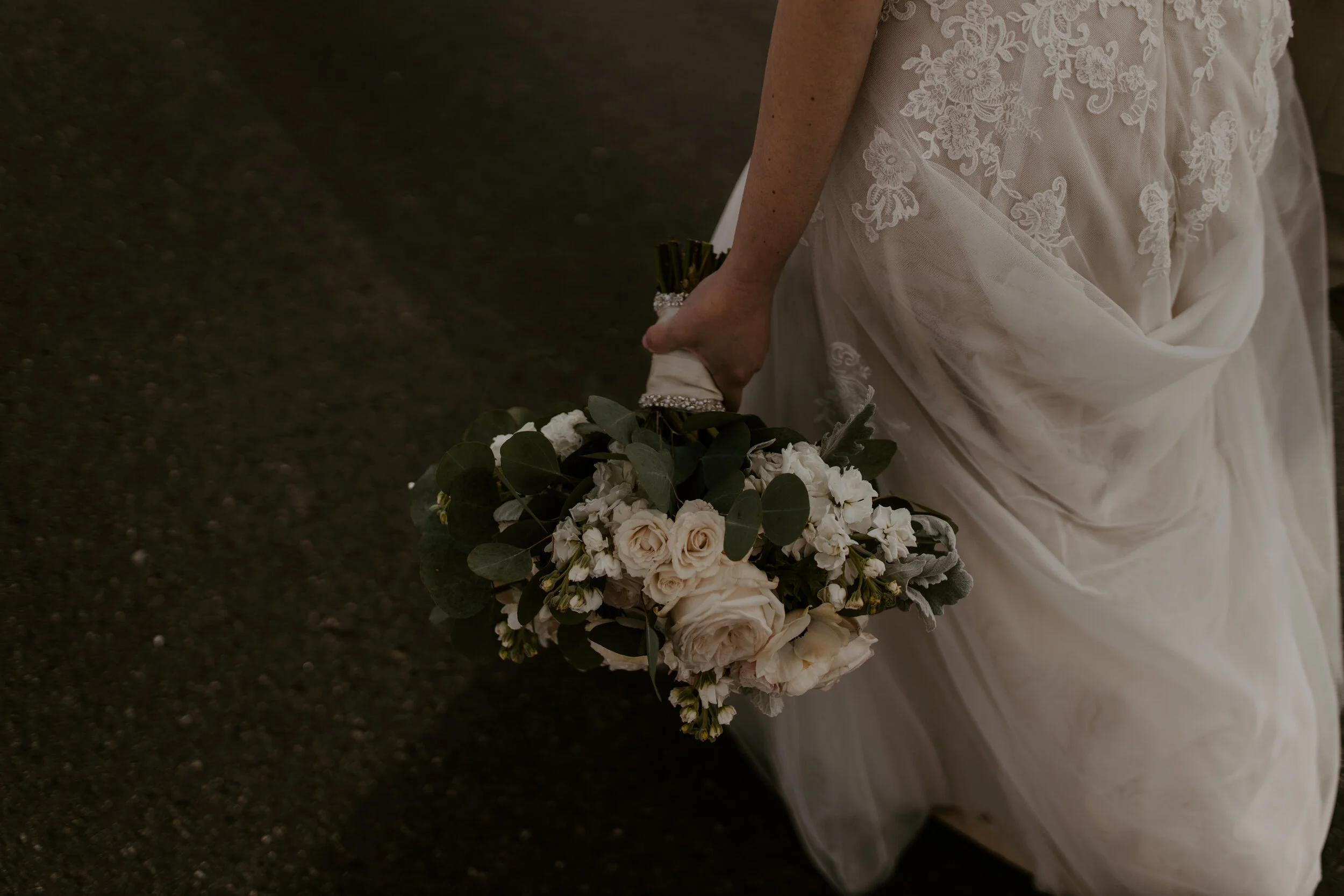 Close-up of a bride in a white lace wedding dress holding a bouquet of white and cream roses and green foliage.