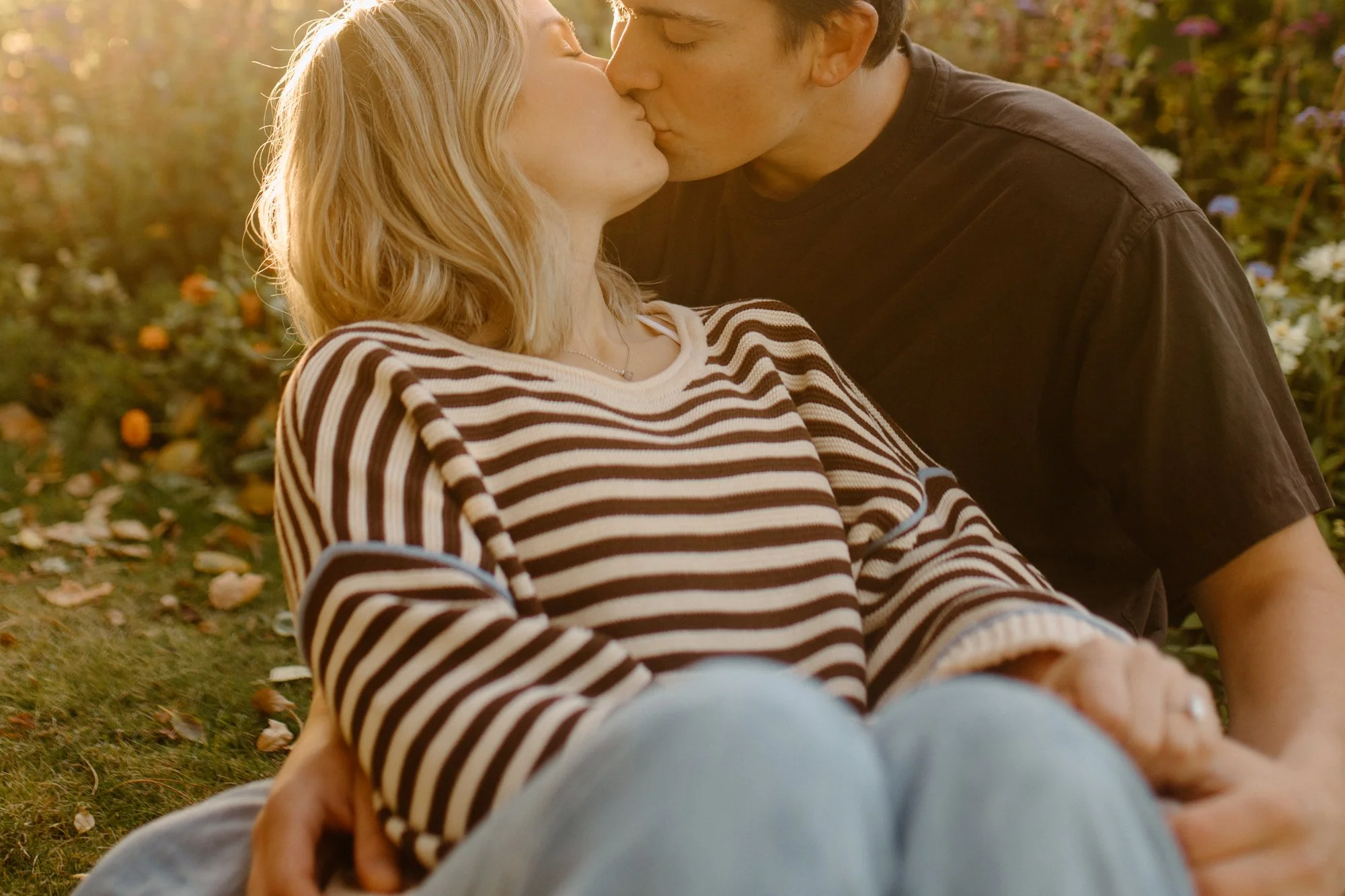 A young couple sharing a kiss outdoors in a garden or park setting during the late afternoon or early evening, with sunlight filtering through the trees, surrounded by flowers and fallen leaves, both sitting on the ground.