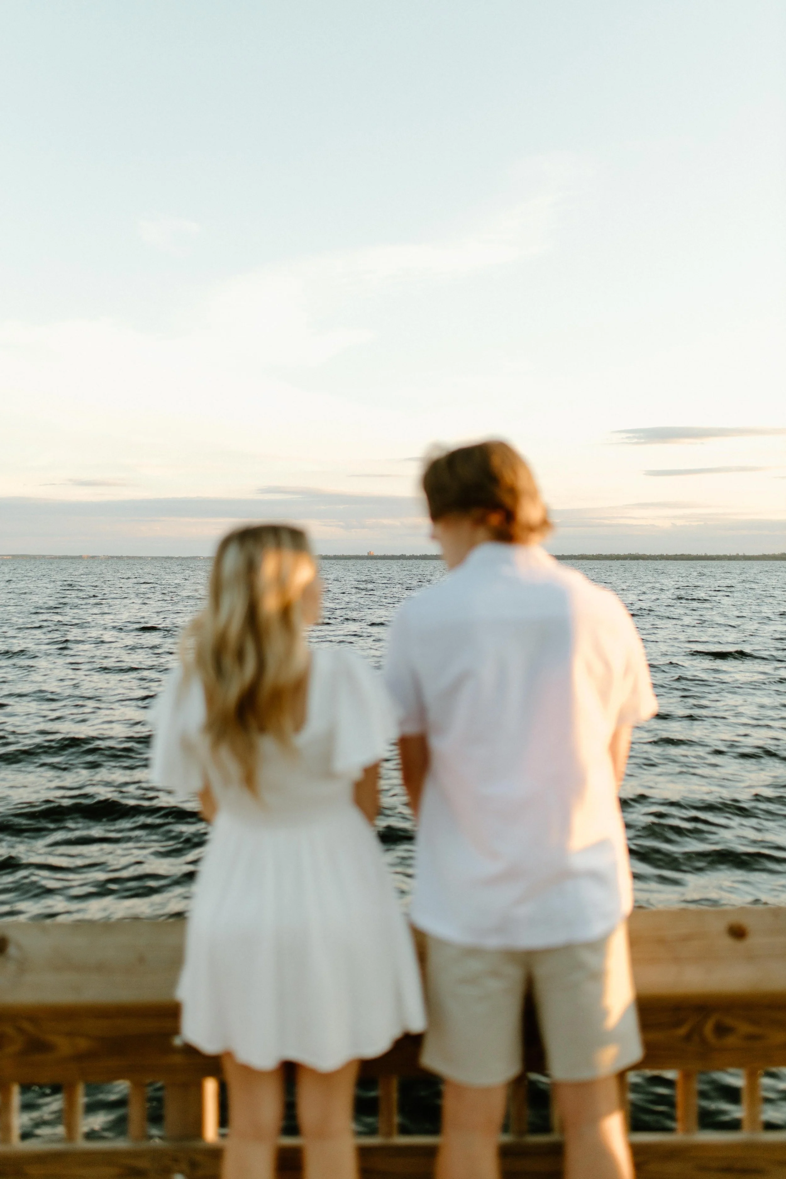 Two young people, a man and a woman, stand on a wooden dock overlooking a large body of water at sunset, with a partly cloudy sky.