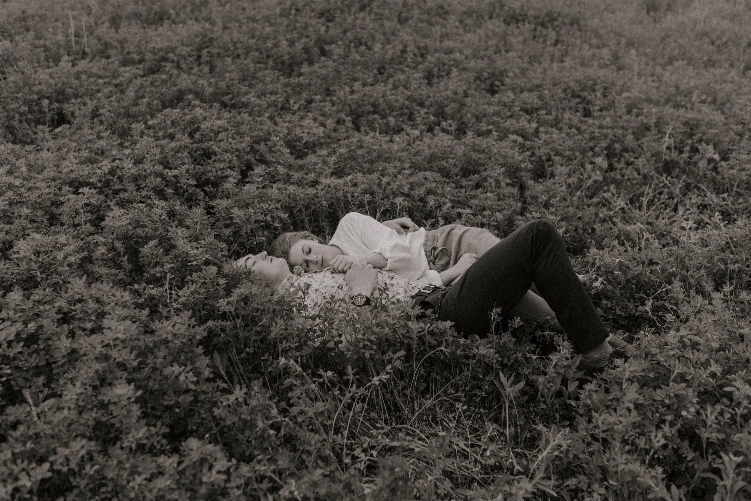 Two people lying in a field of dense low vegetation, resting or sleeping with eyes closed.