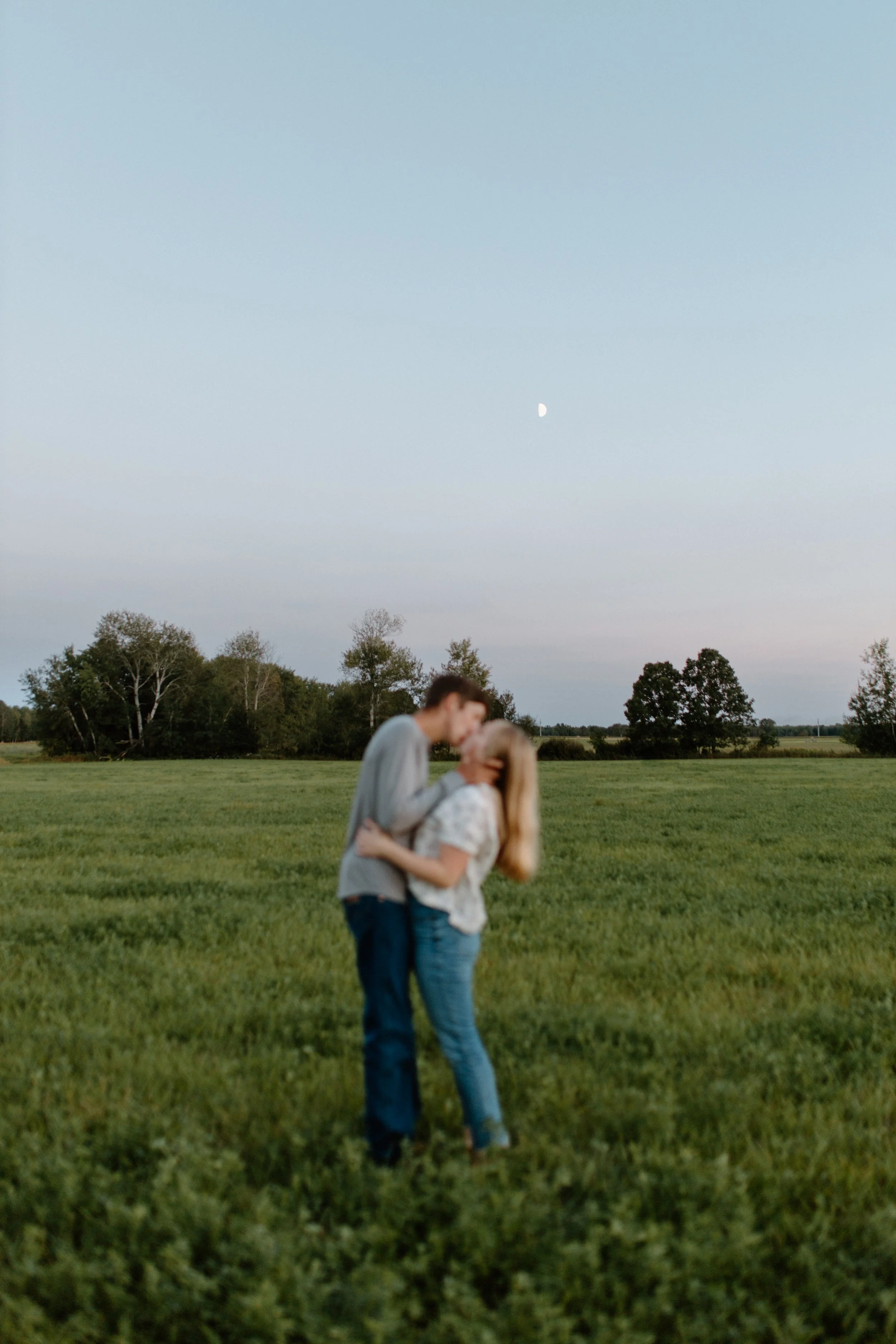 A couple standing outside on a grassy field, embracing and kissing, with trees in the background. The sky is clear with a visible moon.