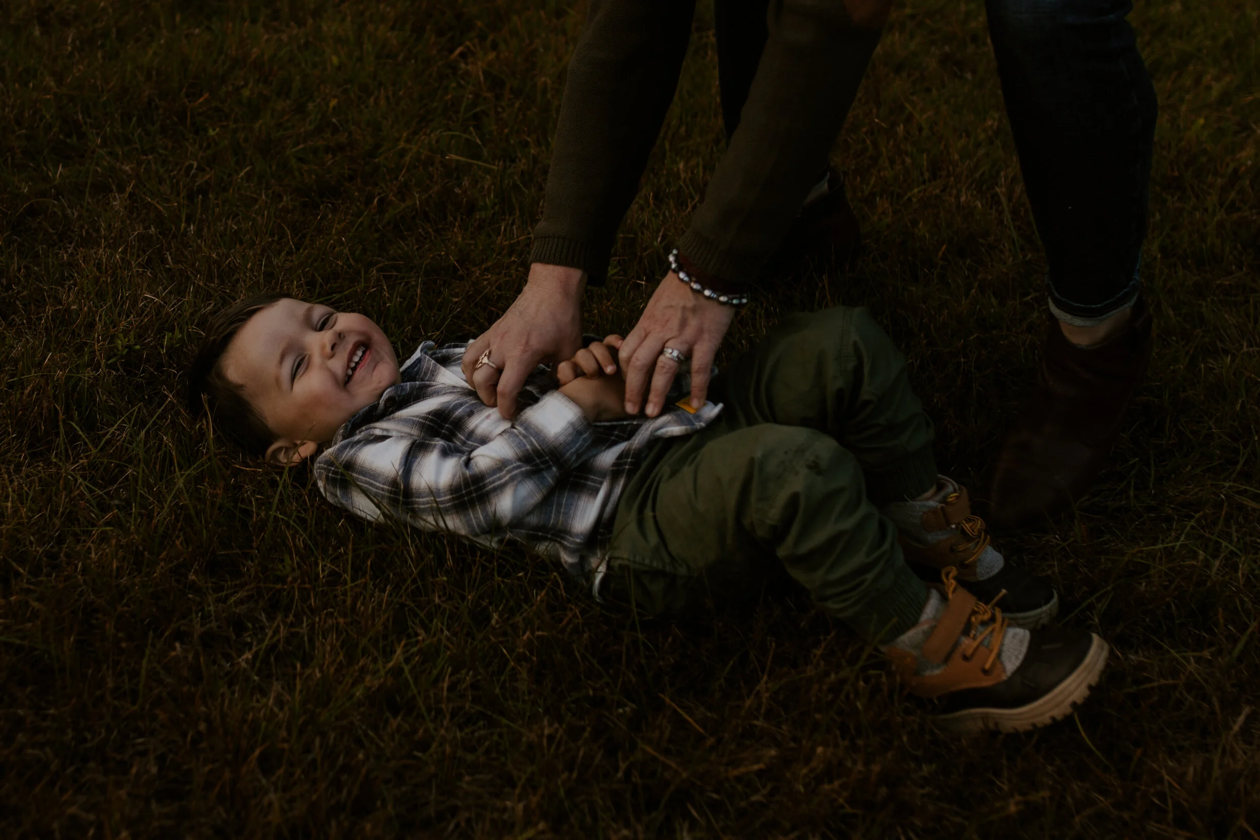 A young boy lying on the grass, smiling, as an adult hands him a snack or toy. The scene is outdoors, possibly during dusk or night.