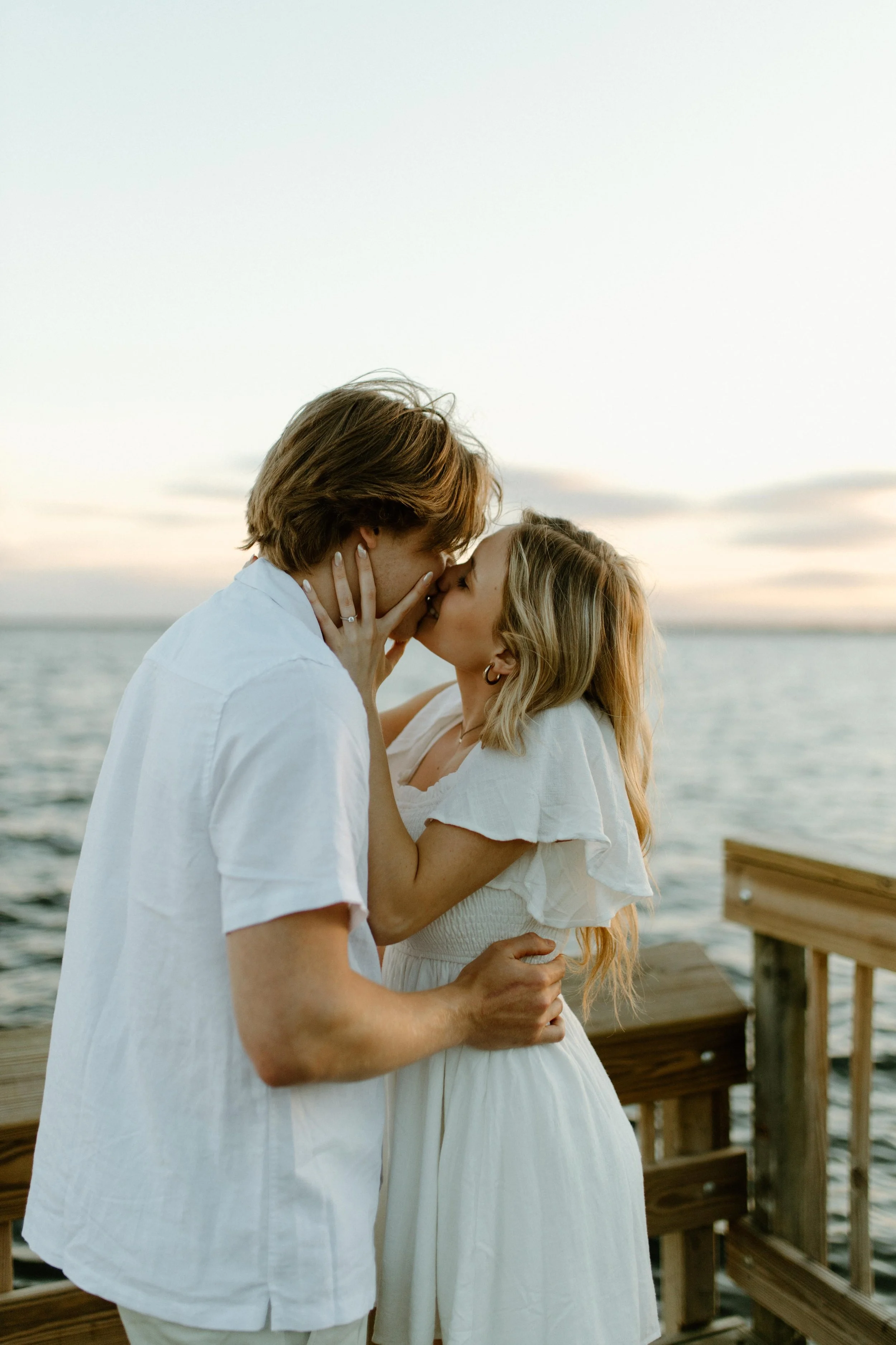 A couple sharing a kiss on a wooden pier by the water during sunset, dressed in white.