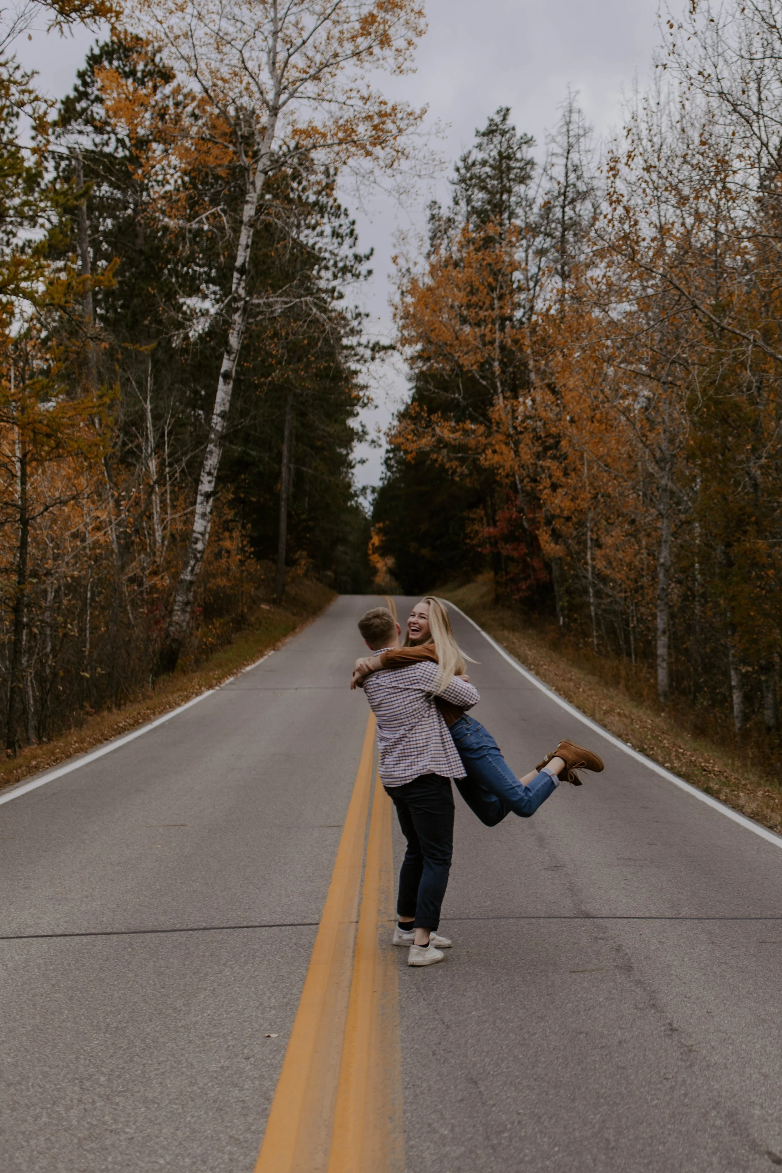 A woman and a man are standing in the middle of a two-lane road, hugging each other. The woman is being swung around by the man and is smiling joyfully. The road is surrounded by trees with fall foliage, and the sky is cloudy.