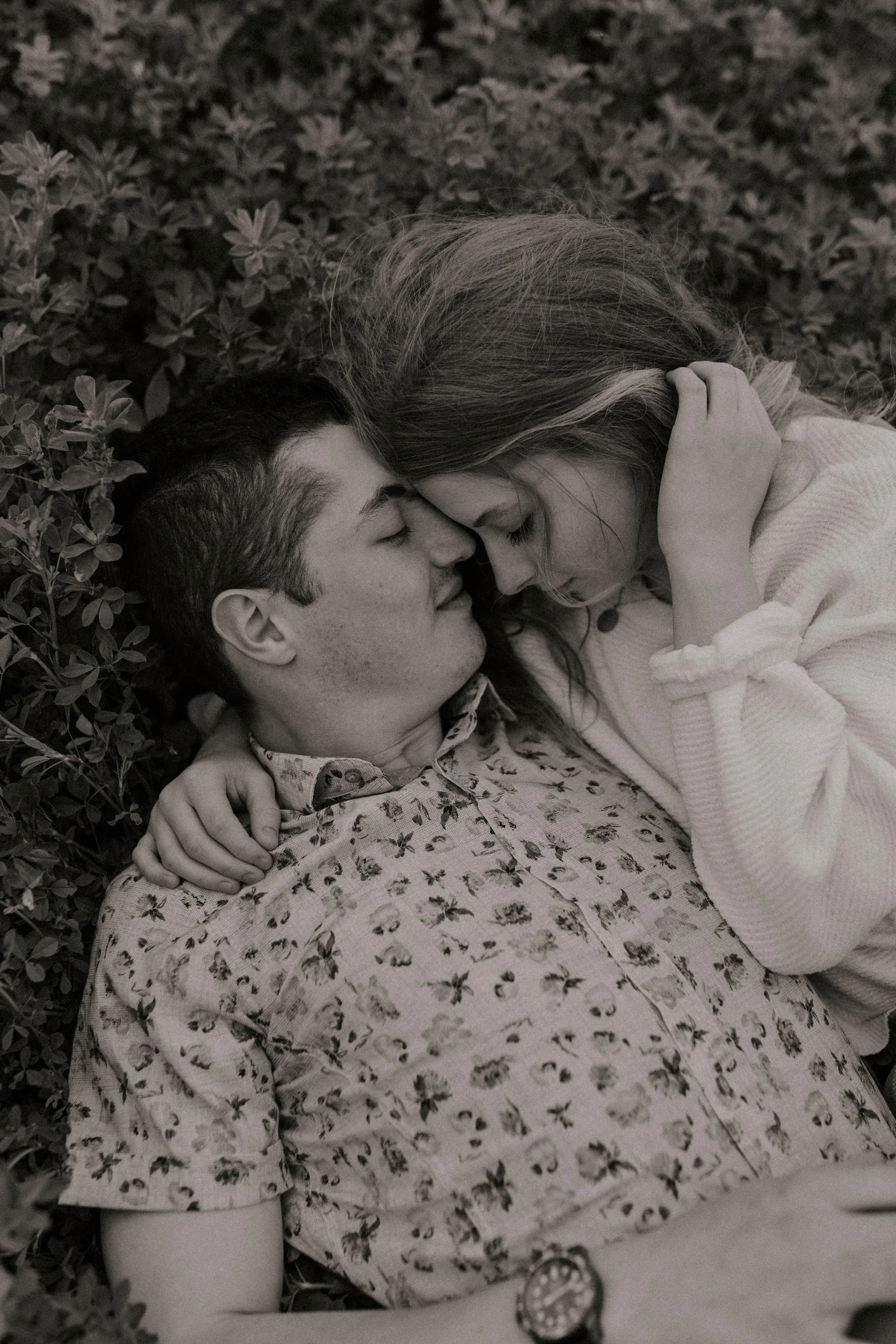A black and white photo of a young couple lying close together with their eyes closed, surrounded by greenery.