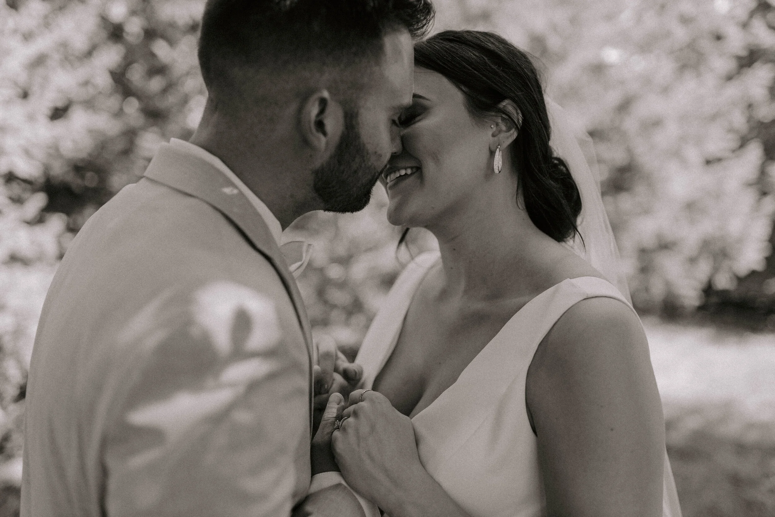 A black-and-white photograph of a couple embracing closely, with their foreheads touching and eyes closed, smiling softly. The background is blurred with what appears to be trees or foliage.