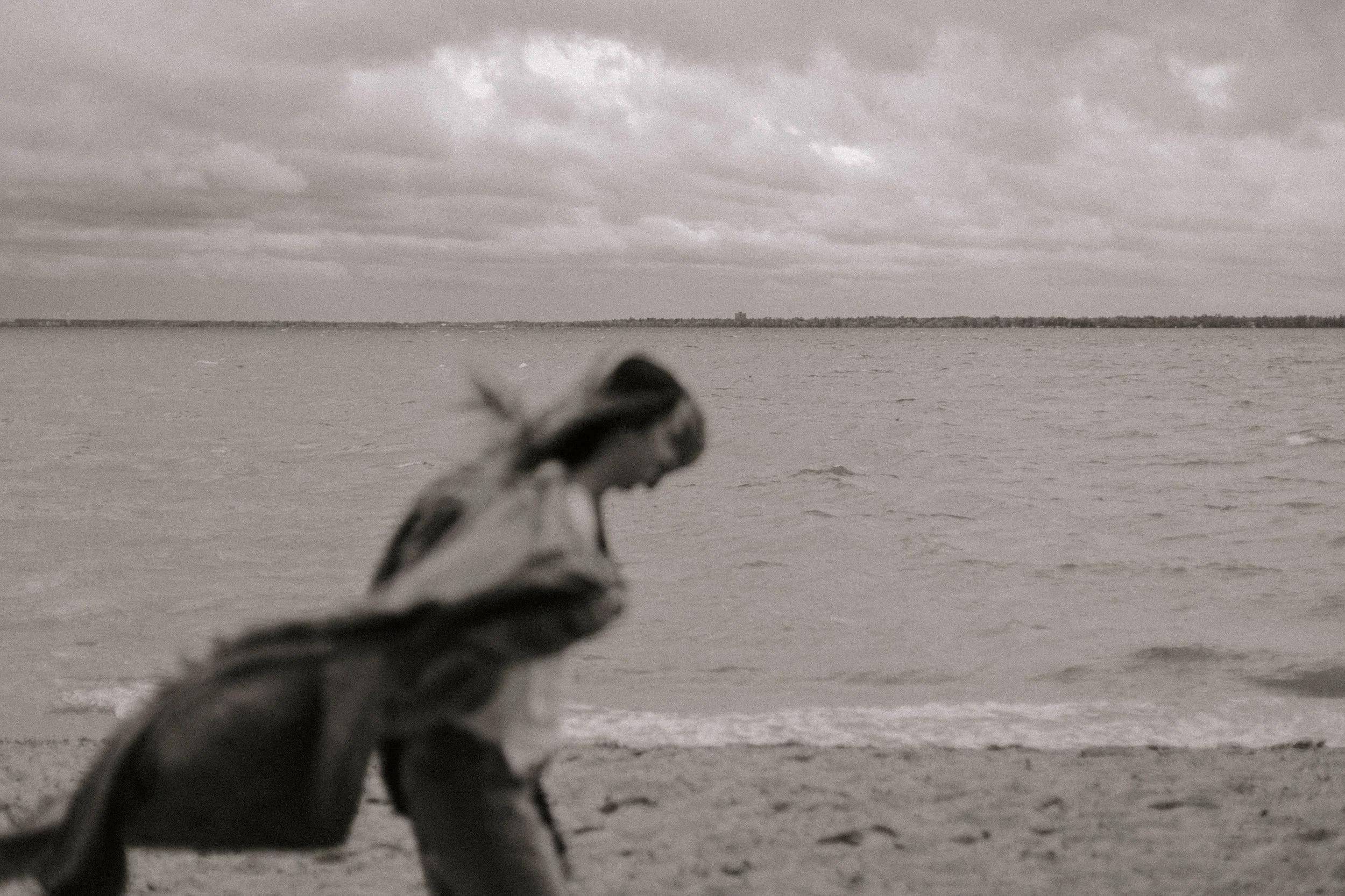 A person lying down on the beach facing the water, with an overcast sky and distant shoreline in the background.