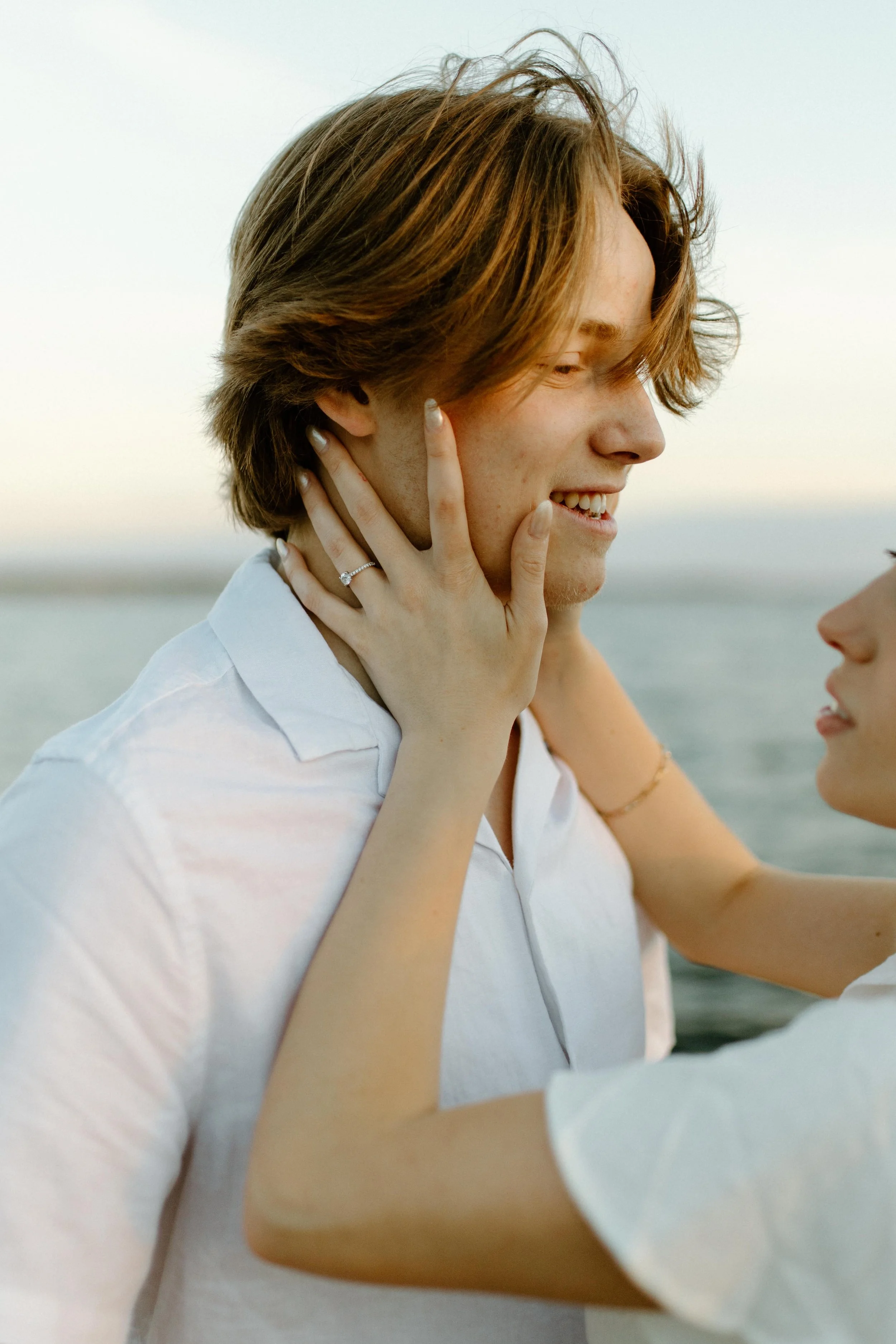 A woman with short brown hair is smiling and holding a person's face, with their hands on her cheeks, against a background of a body of water and a cloudy sky.