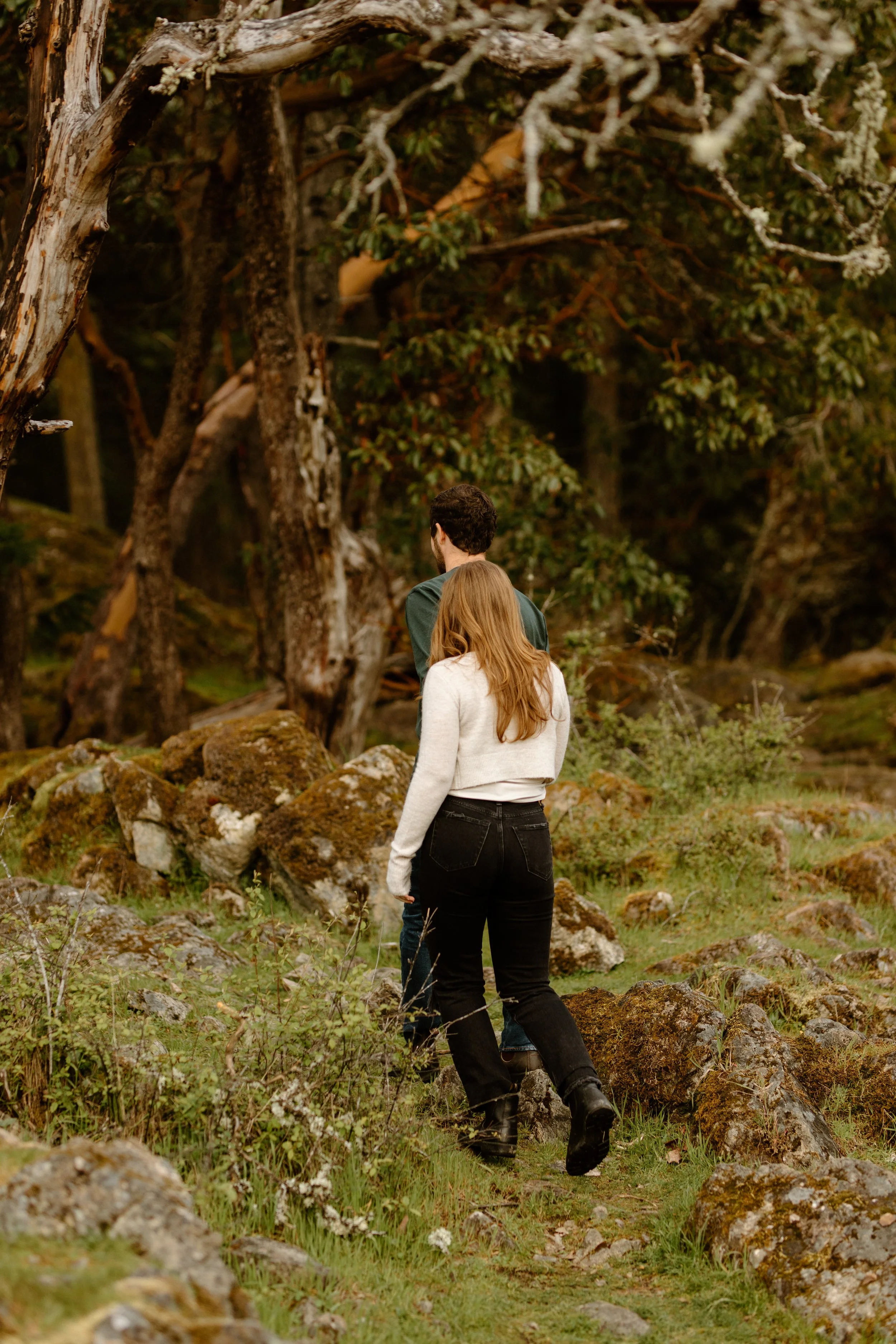 A man and a woman walking through a rocky, forested area with trees and moss-covered rocks.