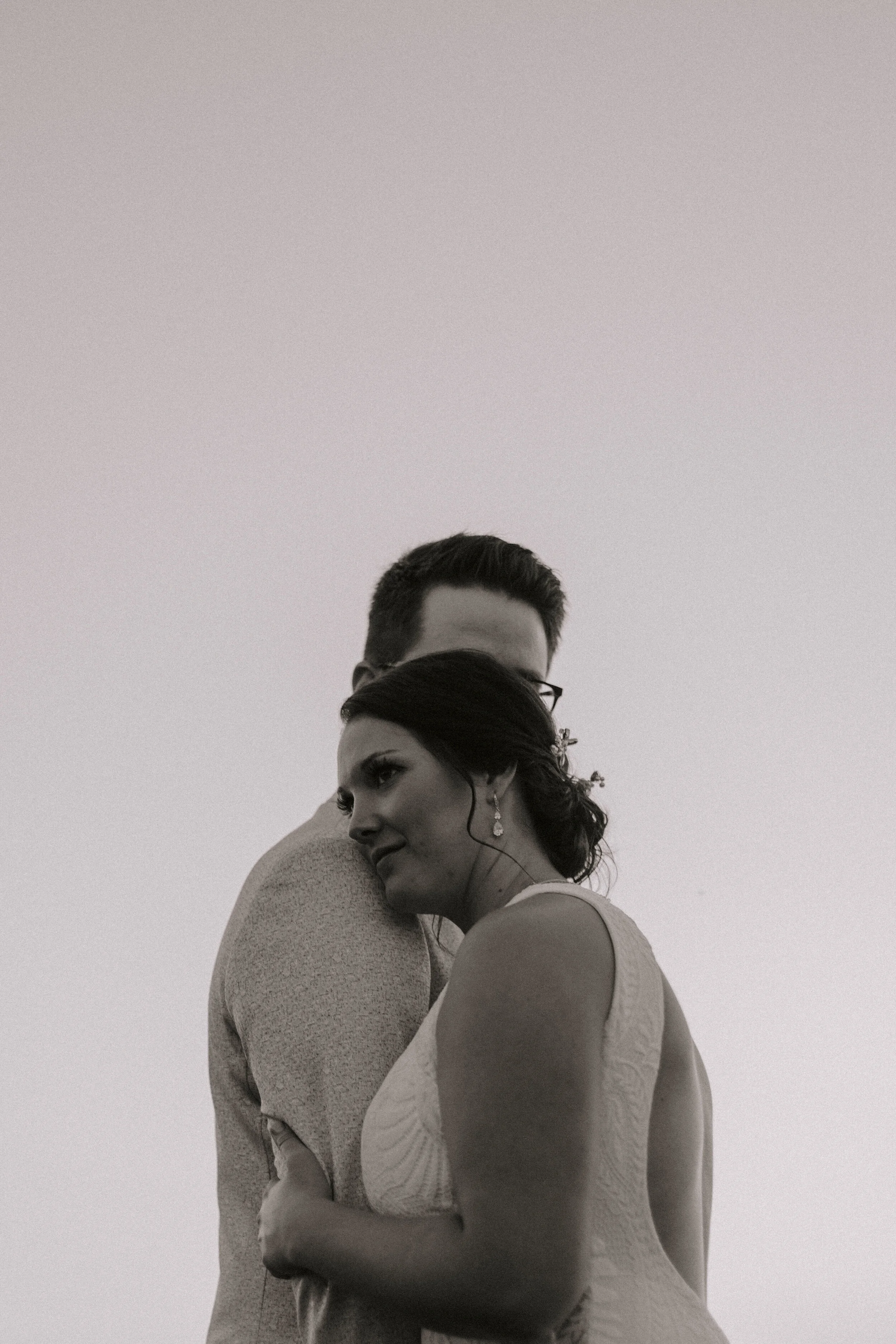 A black and white photo of a woman and man closely embracing, with the woman's eyes closed and a gentle smile, both wearing formal attire, against a plain, light background.