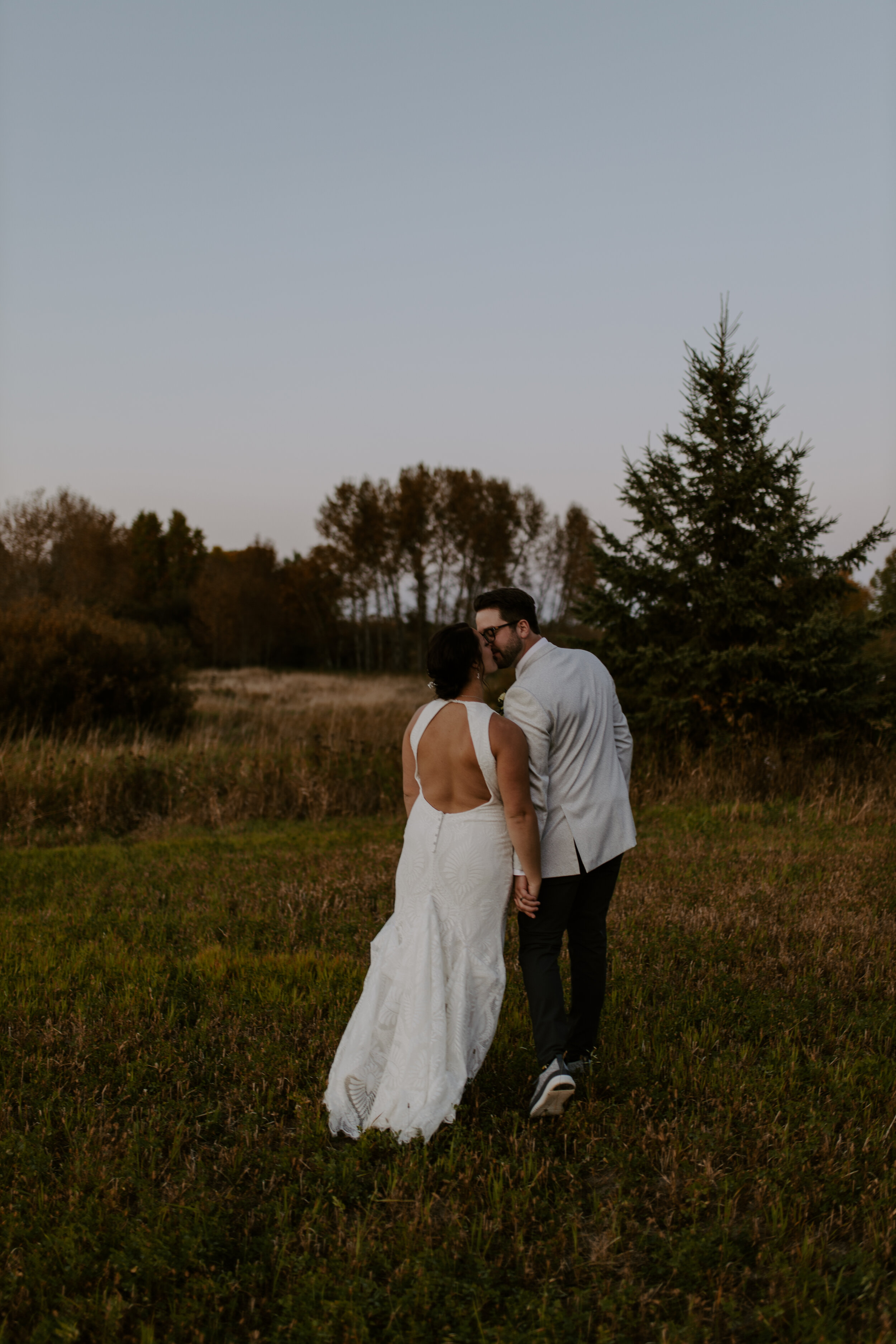 A couple dressed in wedding attire standing close and holding hands in a field with trees in the background during dusk.